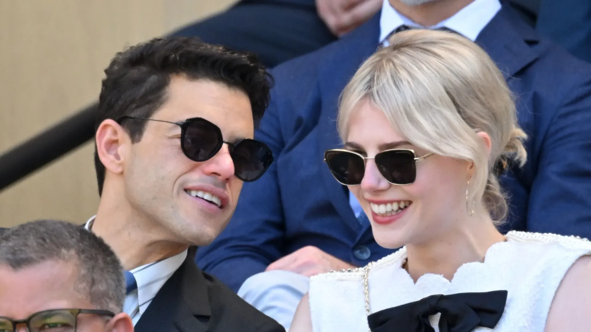 Um homem e uma mulher de óculos de sol estão sentados nas bancadas do estádio