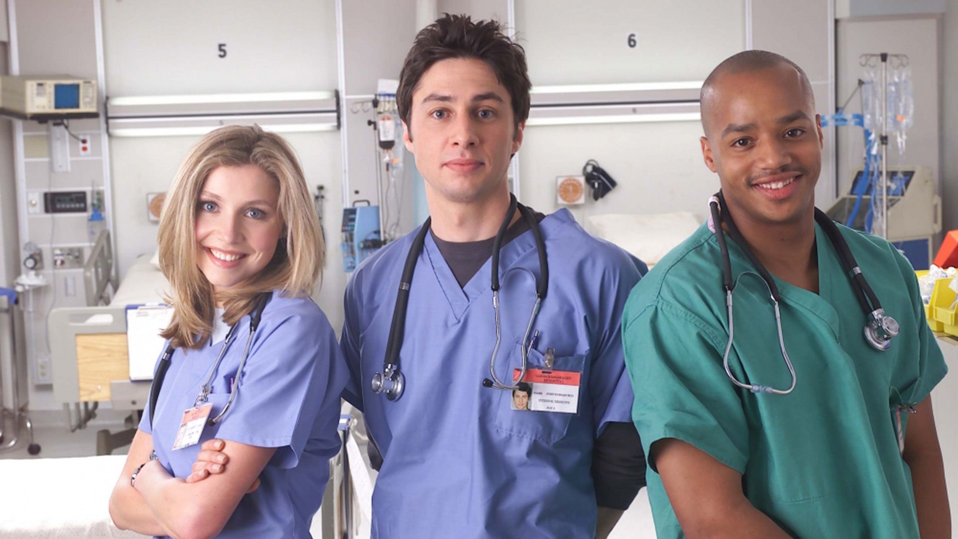 Sarah Chalke, Zach Braff e Donald Faison com batas médicas num corredor de hospital a sorrir