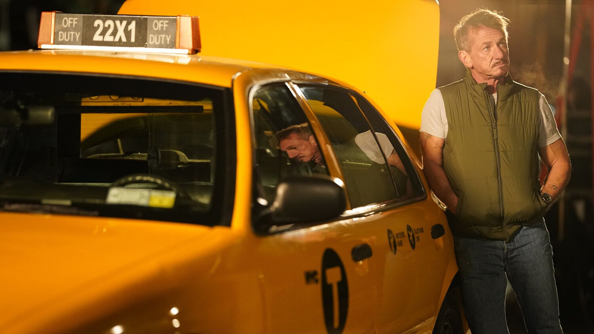 Sean Penn com colete verde encostado a um táxi amarelo