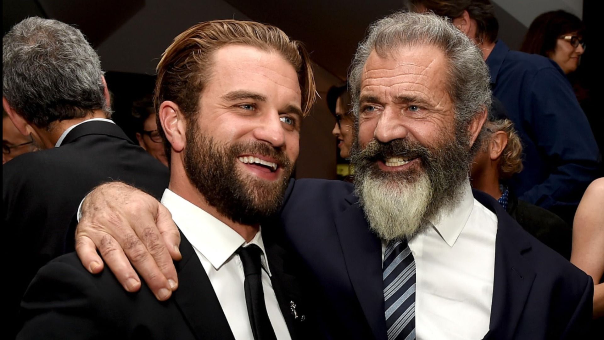 Os atores Milo e Mel Gibson abraçam-se em fatos pretos