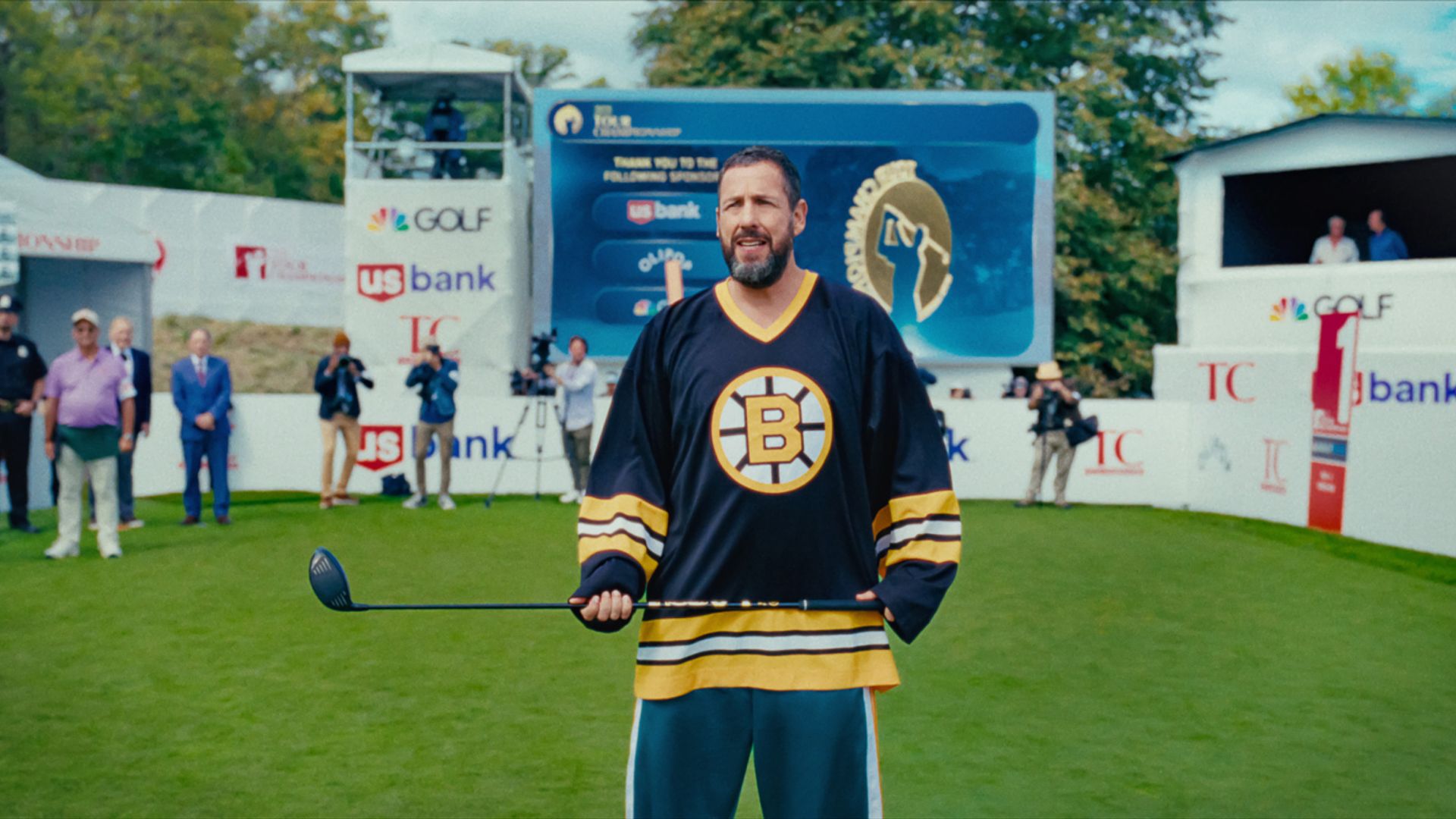 Adam Sandler com uma camisola segura um taco de hóquei enquanto está num estádio