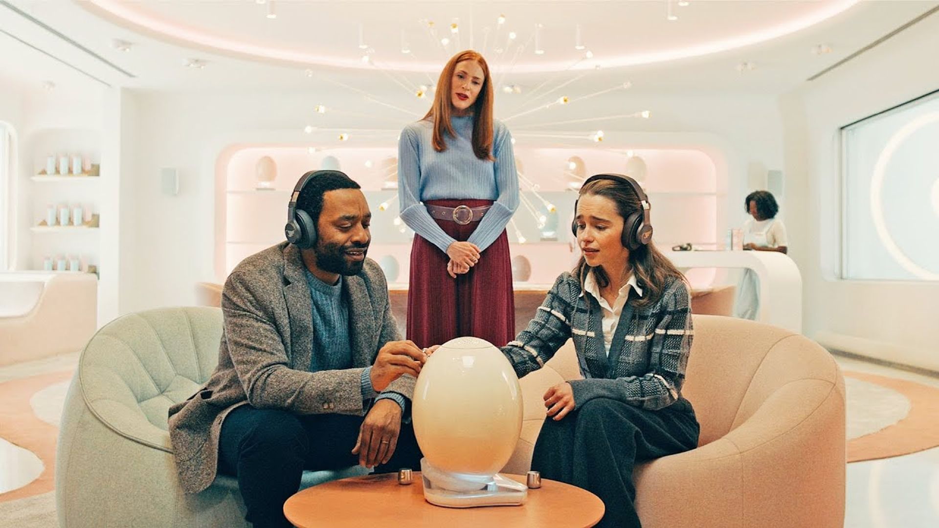 Emilia Clarke e Chiwetel Ejiofor sentados em cadeiras, cenário futurista, cena de The Pod Generation