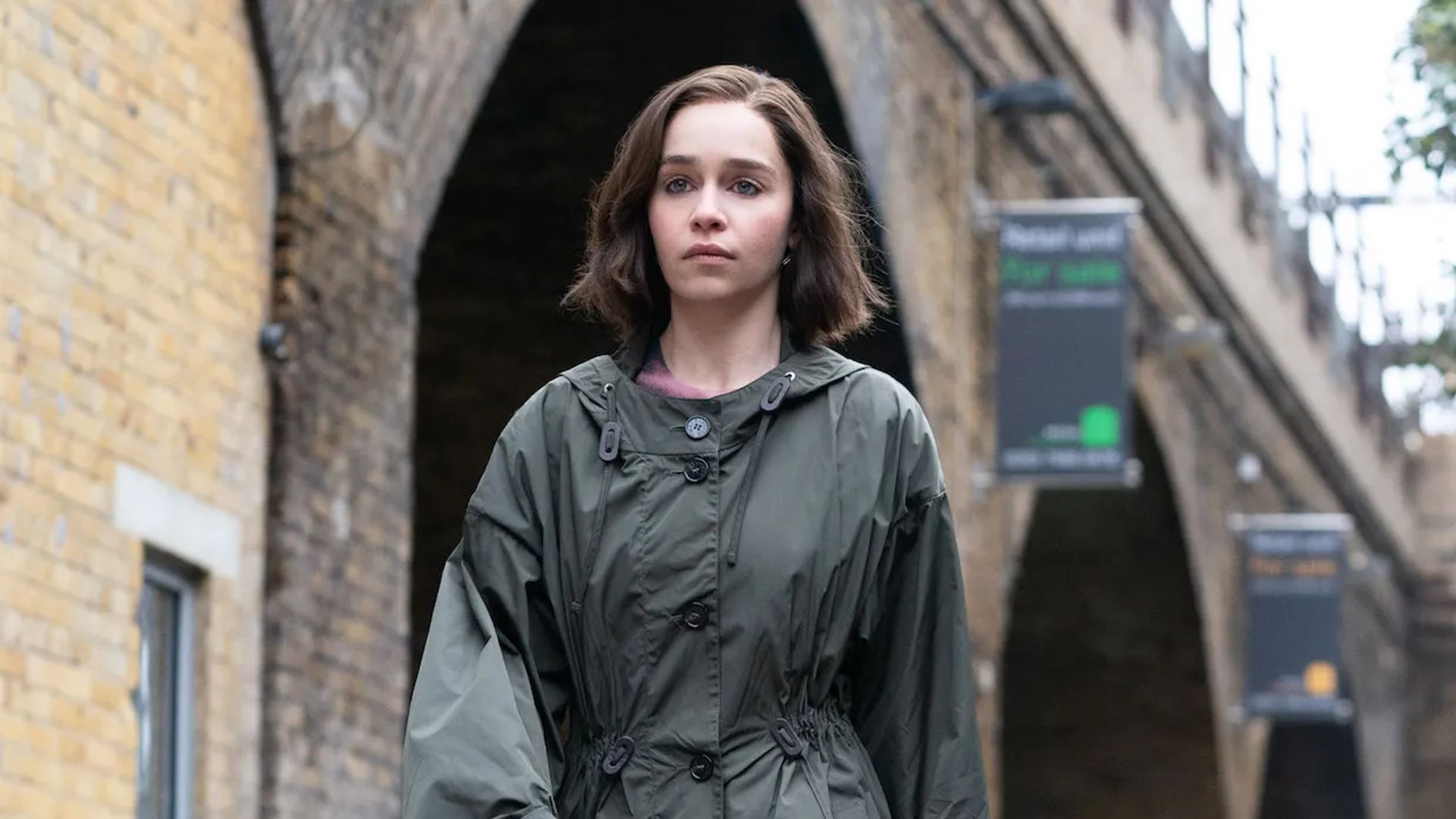Emilia Clarke de capa de chuva caminhando pela rua, olhar focado à frente