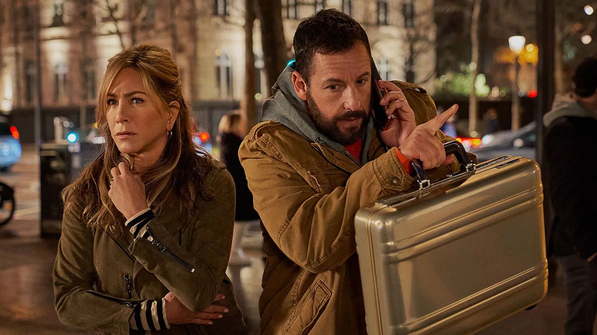 Jennifer Aniston e Adam Sandler numa rua, ela parece assustada, ele está ao telefone e segura uma pasta
