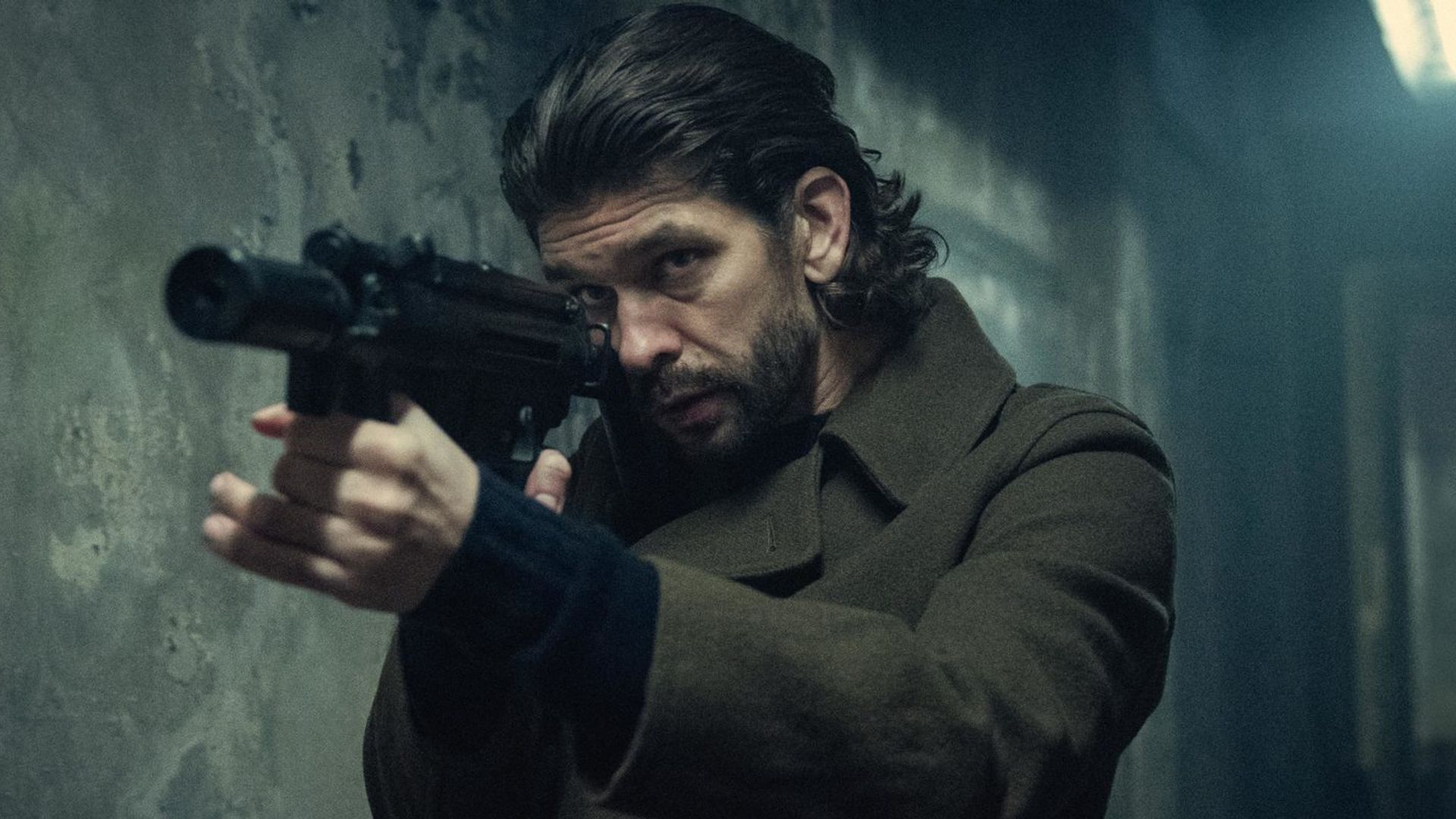 Ben Whishaw de cabelo comprido, de casaco, segurando uma arma