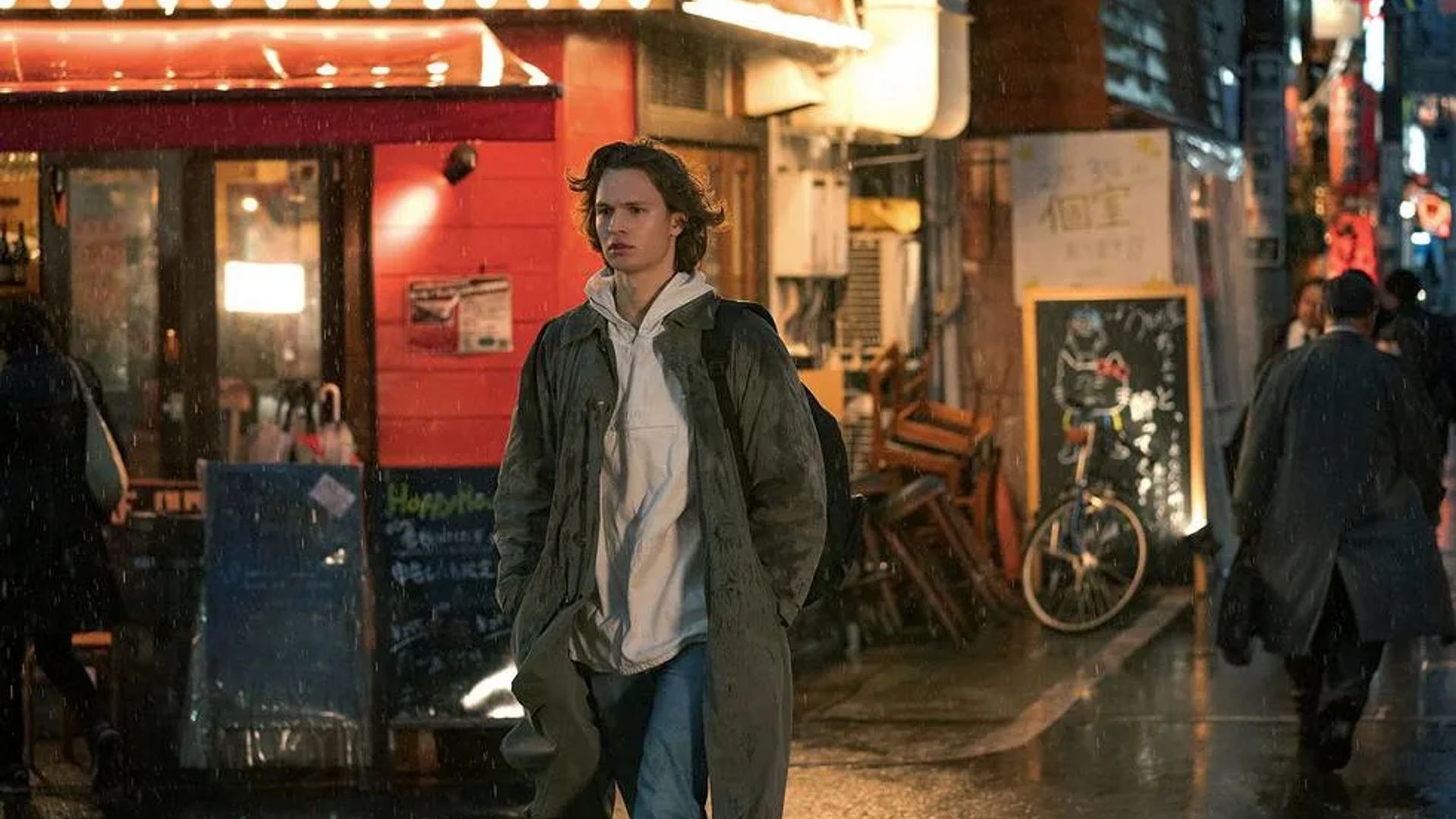 Ansel Elgort de casaco e capuz a caminhar por uma rua chuvosa passando por montras e restaurantes iluminados