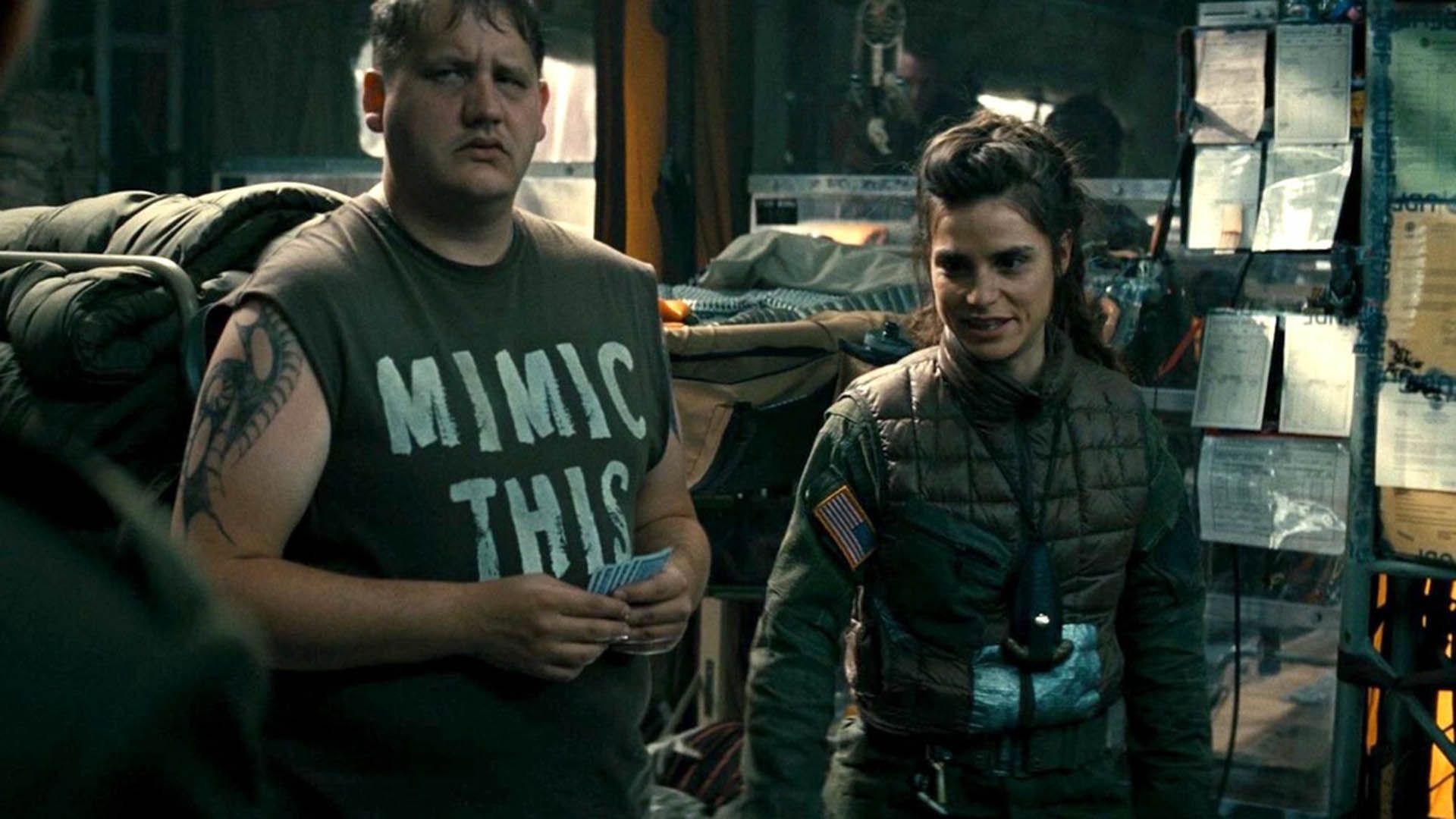 Charlotte Riley em uniforme caqui ao lado de um homem grande numa camisola de alças preta