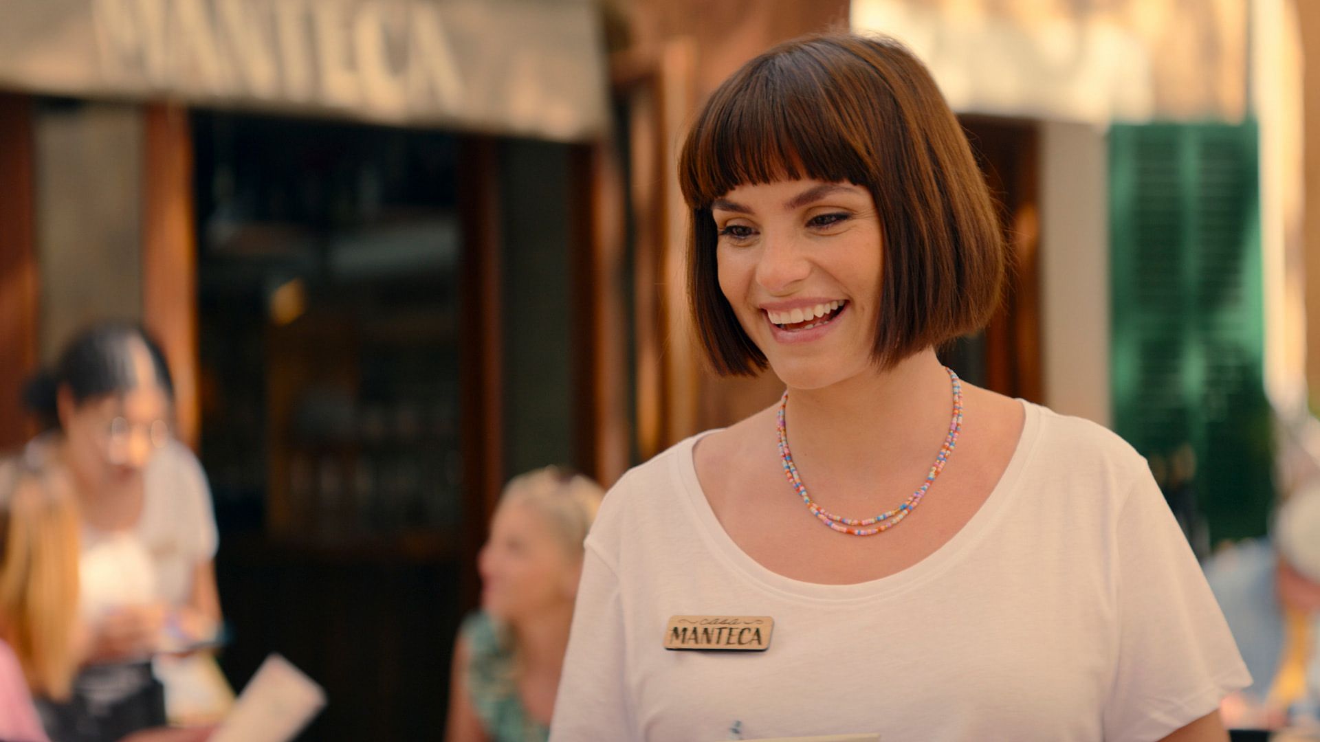 Charlotte Riley com um corte de cabelo bob usando uma camiseta branca, um crachá de madeira no peito e um colar colorido de contas