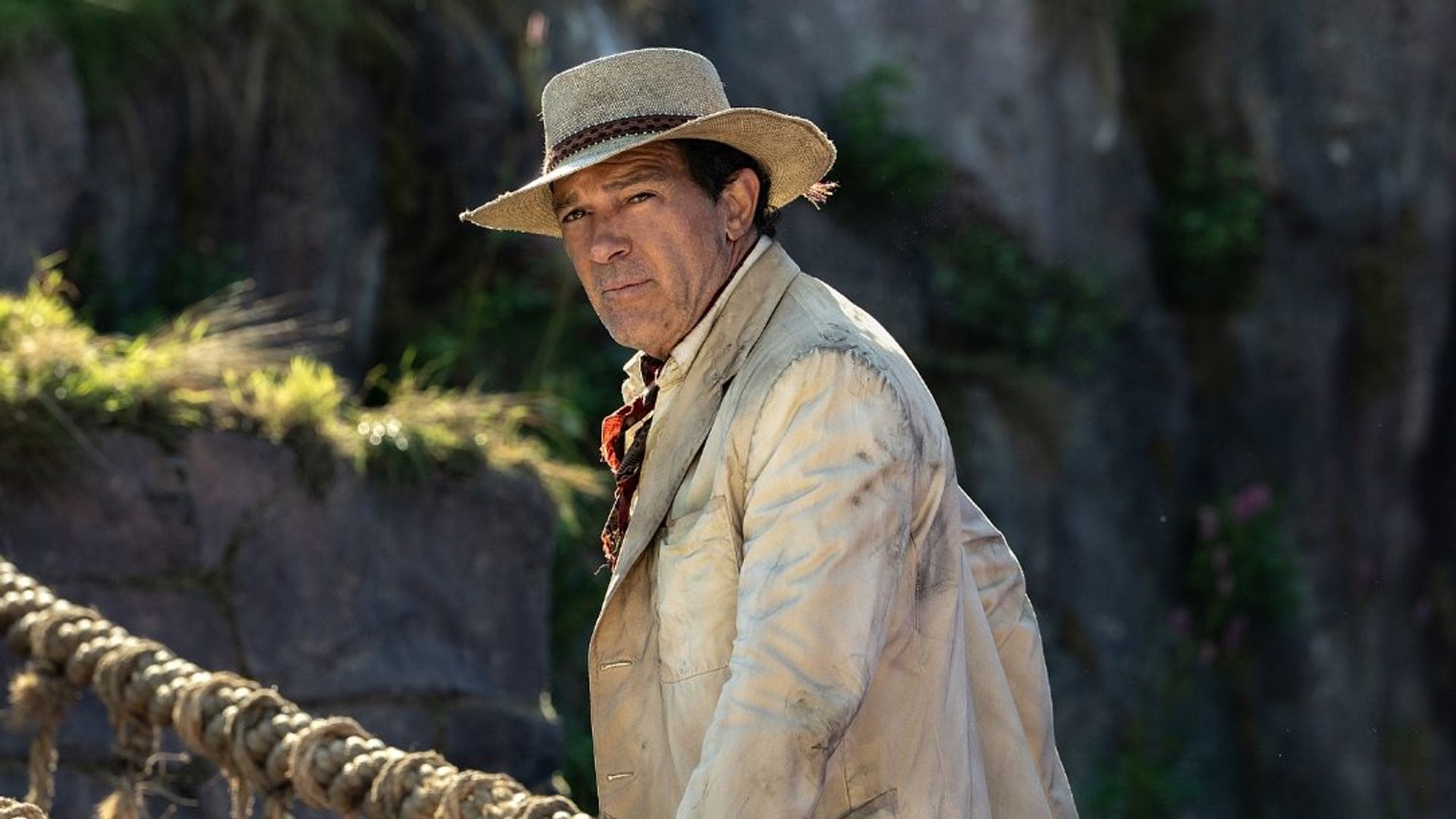 Antonio Banderas em traje retro de aventureiro contra um cenário de selva com ponte suspensa