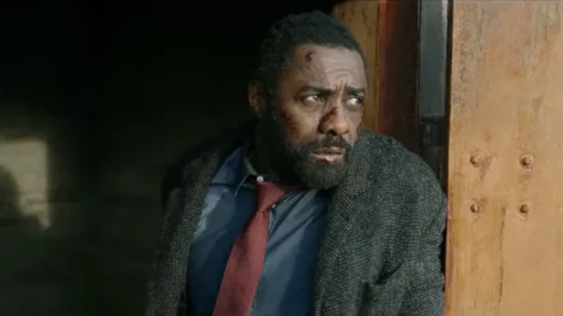 Idris Elba de casaco e gravata vermelha olhando para o lado, still do filme