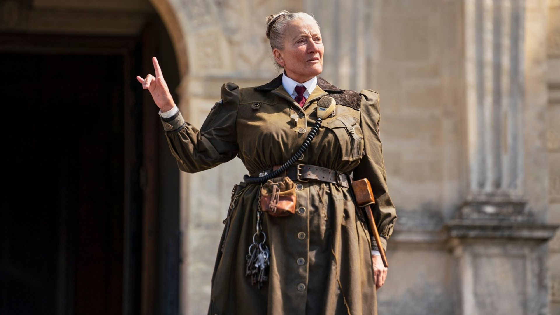 Emma Thompson maquilhada e caracterizada com próteses em frente a um edifício histórico