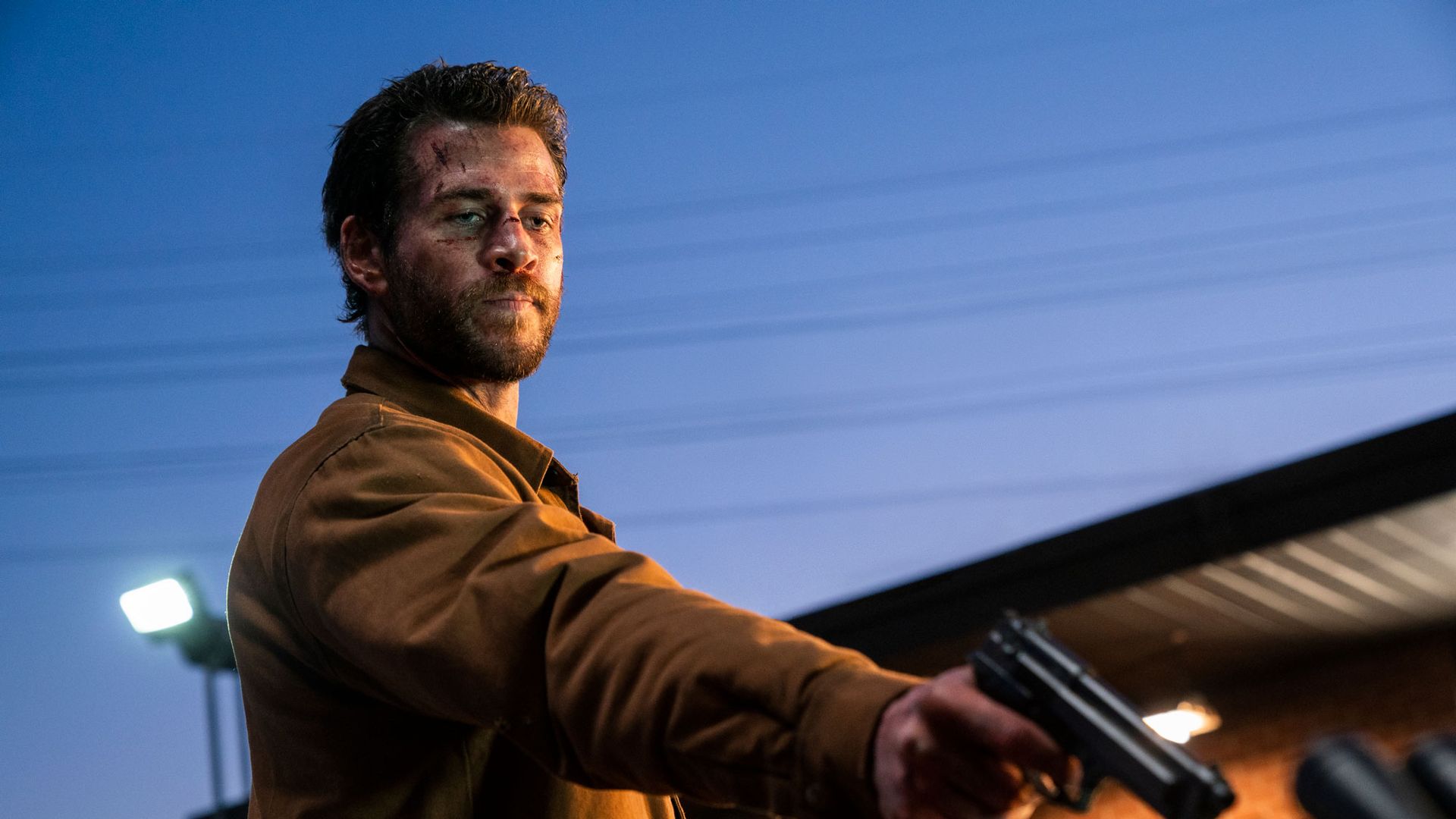 Liam Hemsworth com um casaco castanho a segurar uma arma