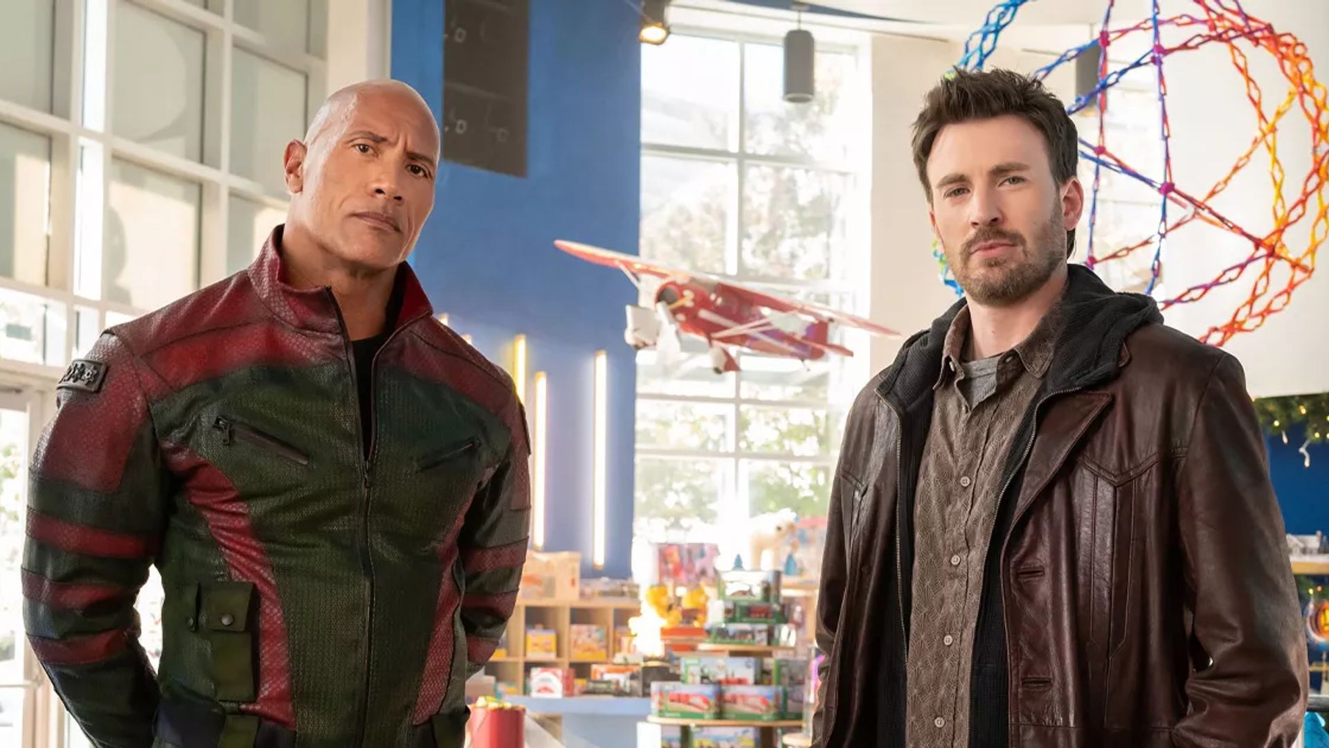 Chris Evans e Dwayne Johnson de casaco numa sala bem iluminada com brinquedos e um modelo de avião ao fundo
