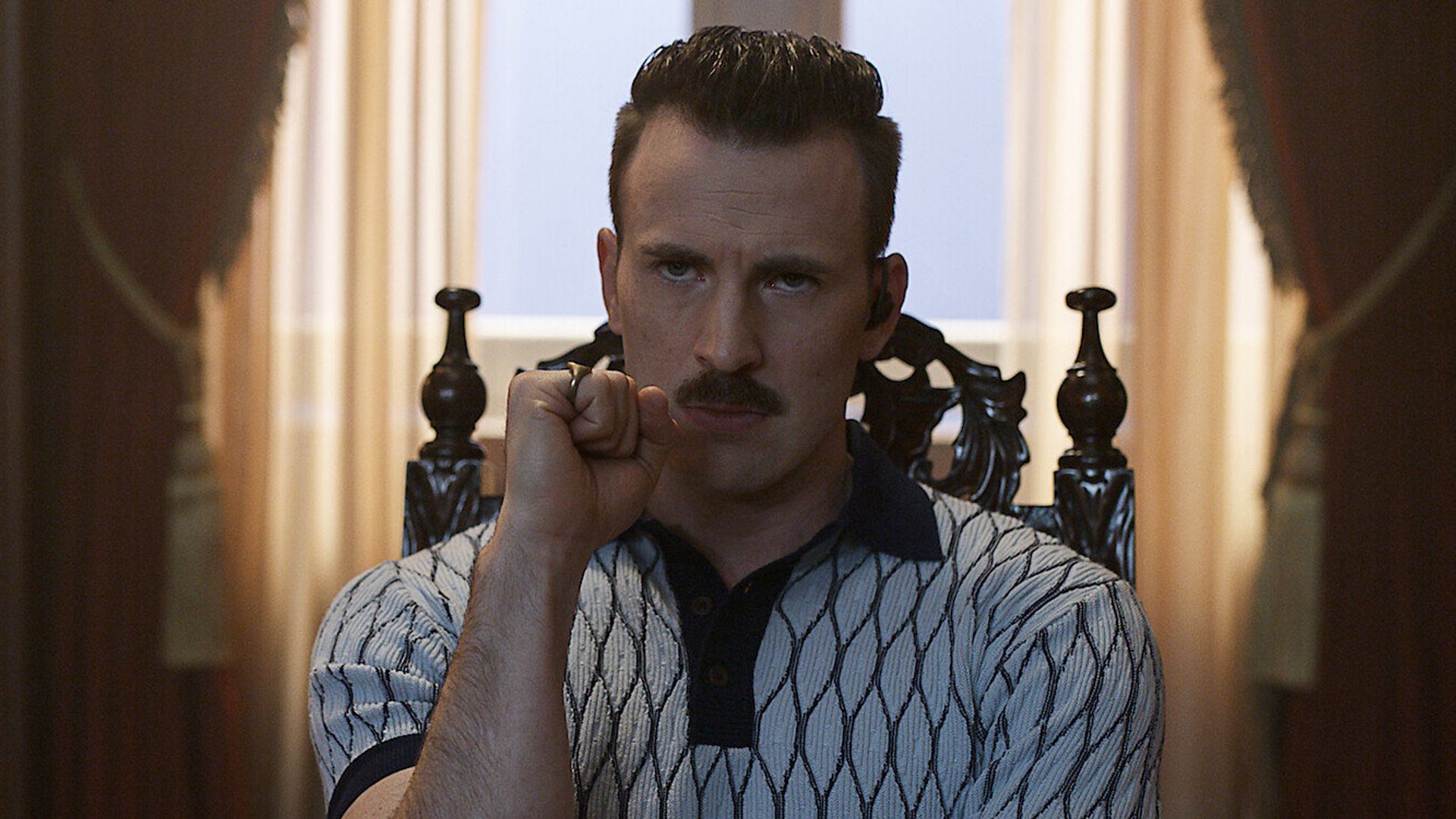 Chris Evans com bigode vestindo uma camisa estampada senta-se numa cadeira ornamentada olhando pensativo para a frente com o punho cerrado contra o rosto