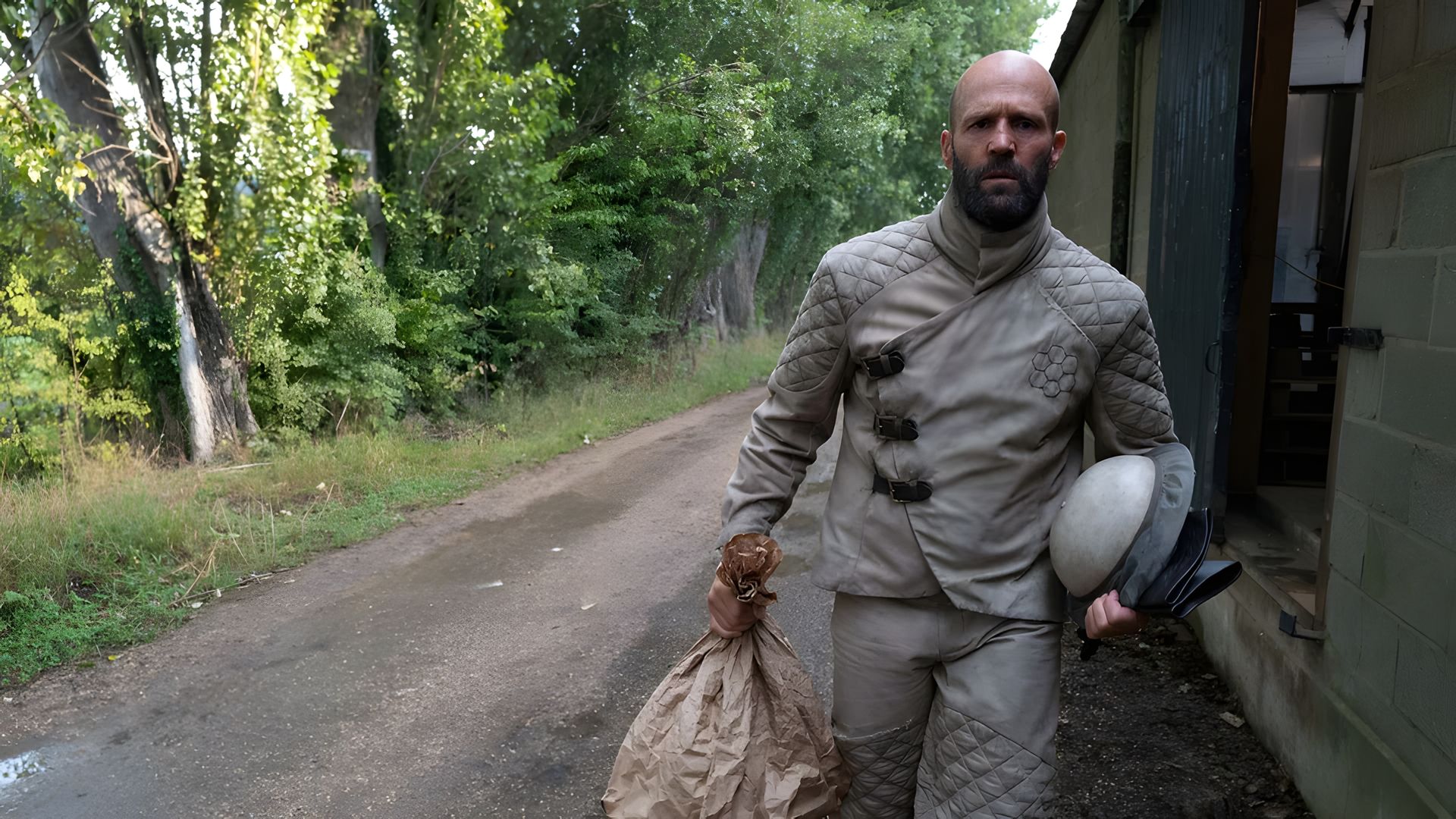 Jason Statham num fato de proteção cinzento a caminhar por uma estrada com um saco e capacete, com árvores ao fundo
