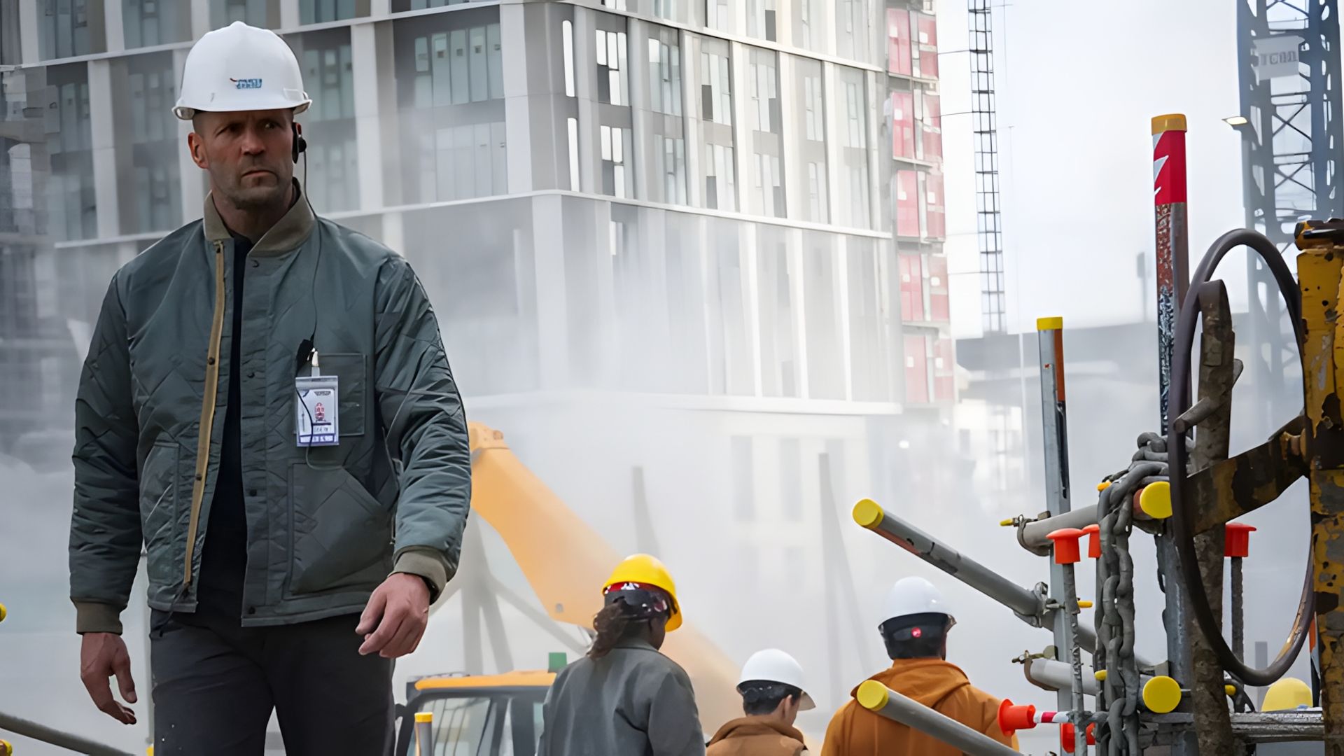 Jason Statham com capacete e casaco caminha por uma obra com trabalhadores e maquinaria visíveis ao fundo enevoado