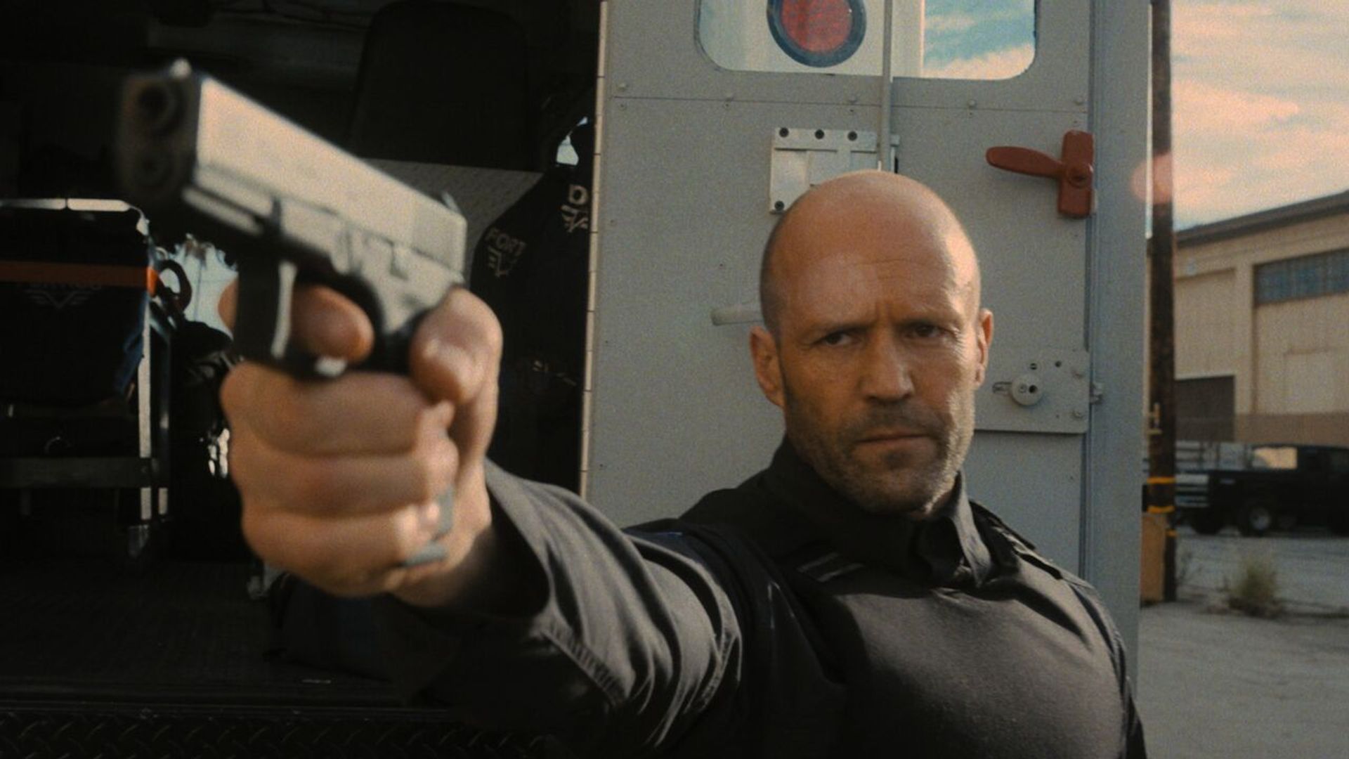 Jason Statham com equipamento tático preto aponta uma pistola junto a um carro-forte contra um cenário industrial