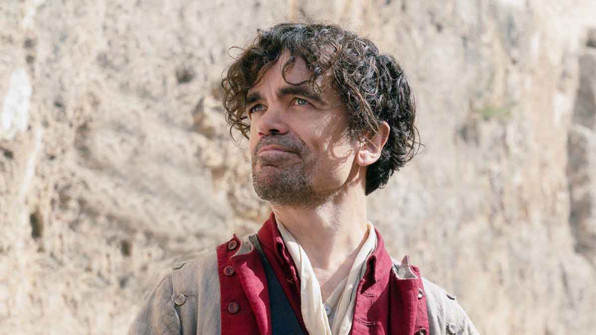 Peter Dinklage em traje de época com gibão vermelho segura uma carta e olha para cima com um ligeiro sorriso