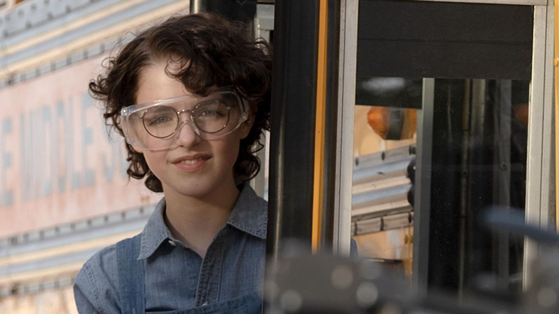 Mckenna Grace com caracóis pretos curtos e óculos, cena do filme