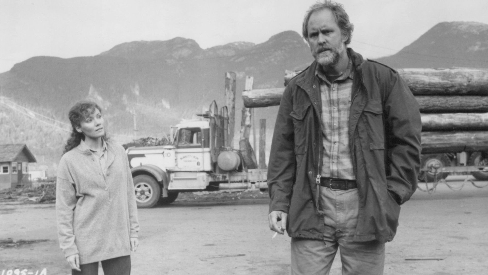 John Lithgow com roupas esfarrapadas contra o cenário de um camião madeireiro numa cena do filme "Distant Thunder"