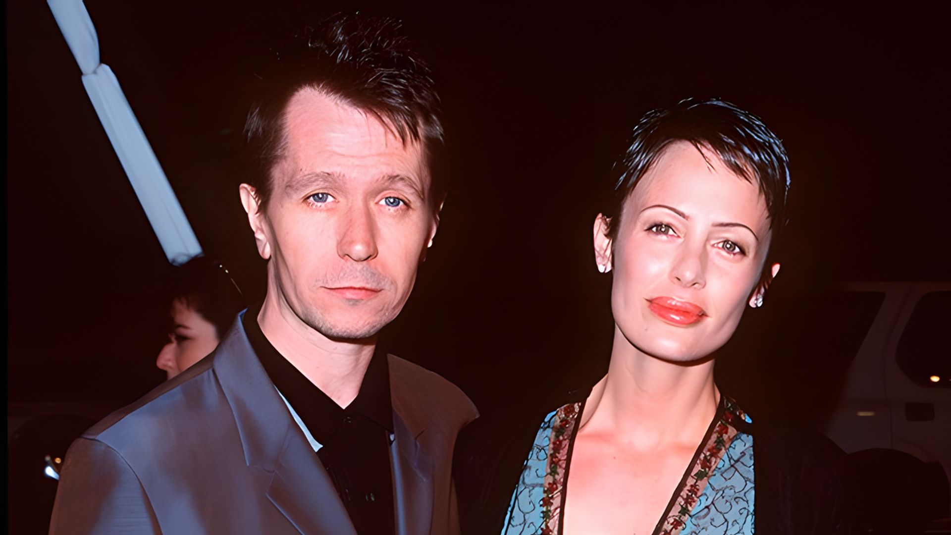 Gary Oldman de fato escuro e Donya Fiorentino num vestido elegante posam juntos num evento noturno.