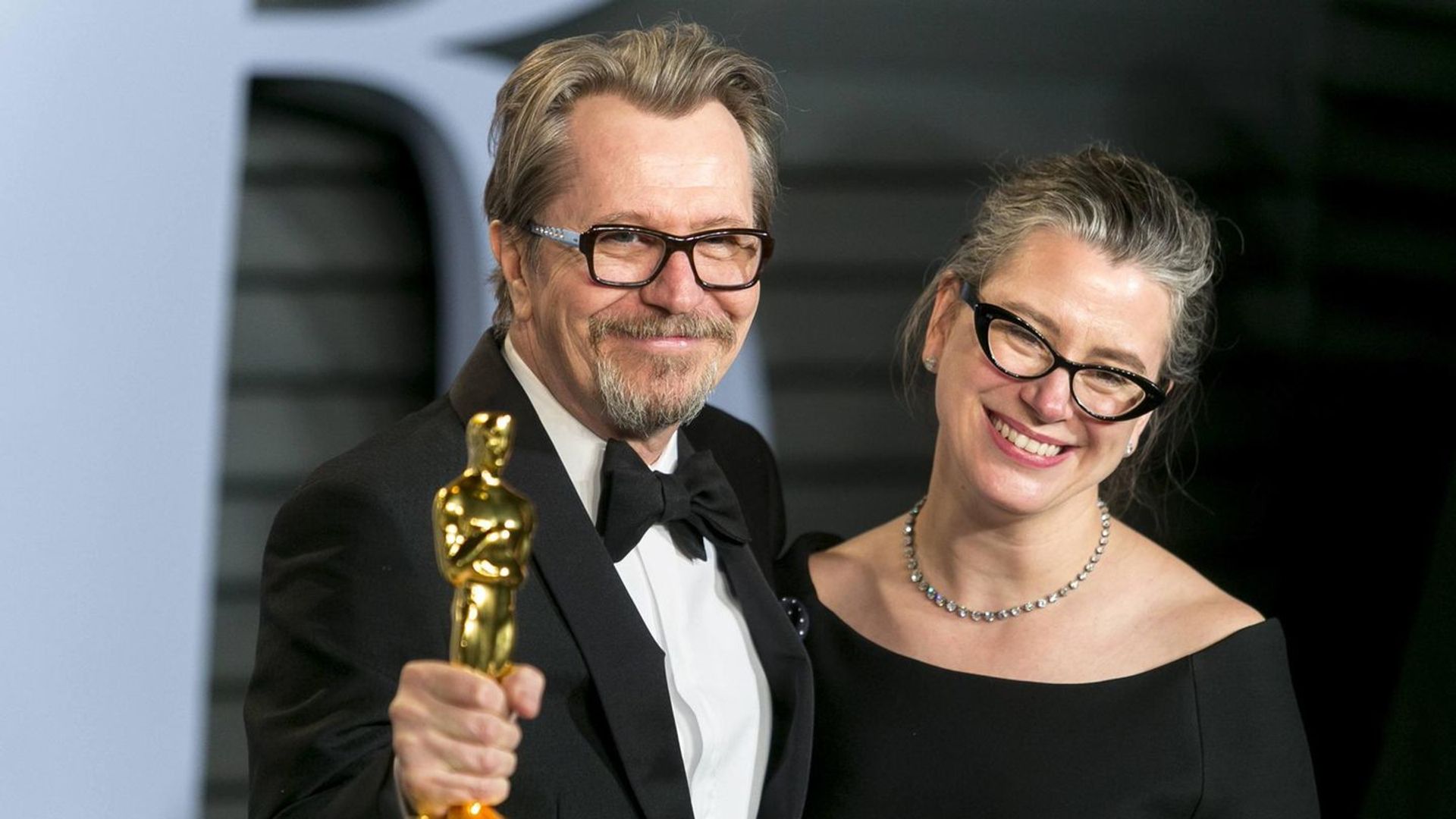 Gary Oldman de smoking segura uma estatueta do Oscar e sorri ao lado de Gisele Schmidt, vestida de preto e usando óculos