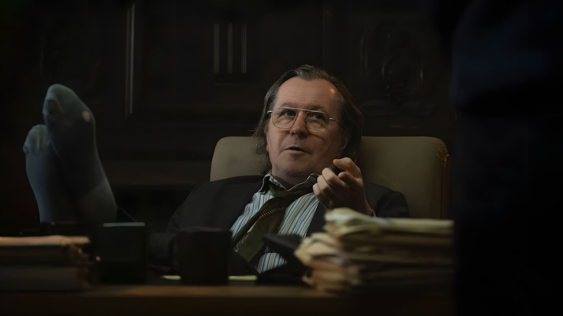 Gary Oldman de óculos e fato sentado numa secretária num escritório com os pés em cima da mesa, com uma expressão pensativa
