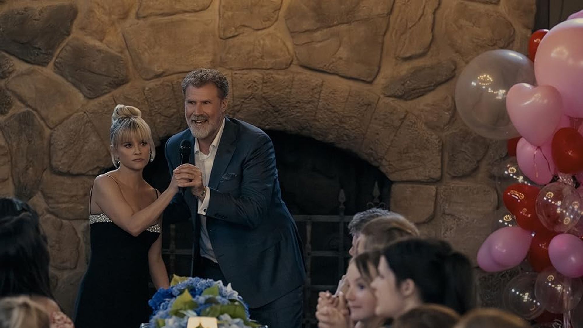 Will Ferrell de fato com um microfone dá as mãos a Reese Witherspoon num vestido preto contra o cenário de uma lareira, mesa festiva e balões