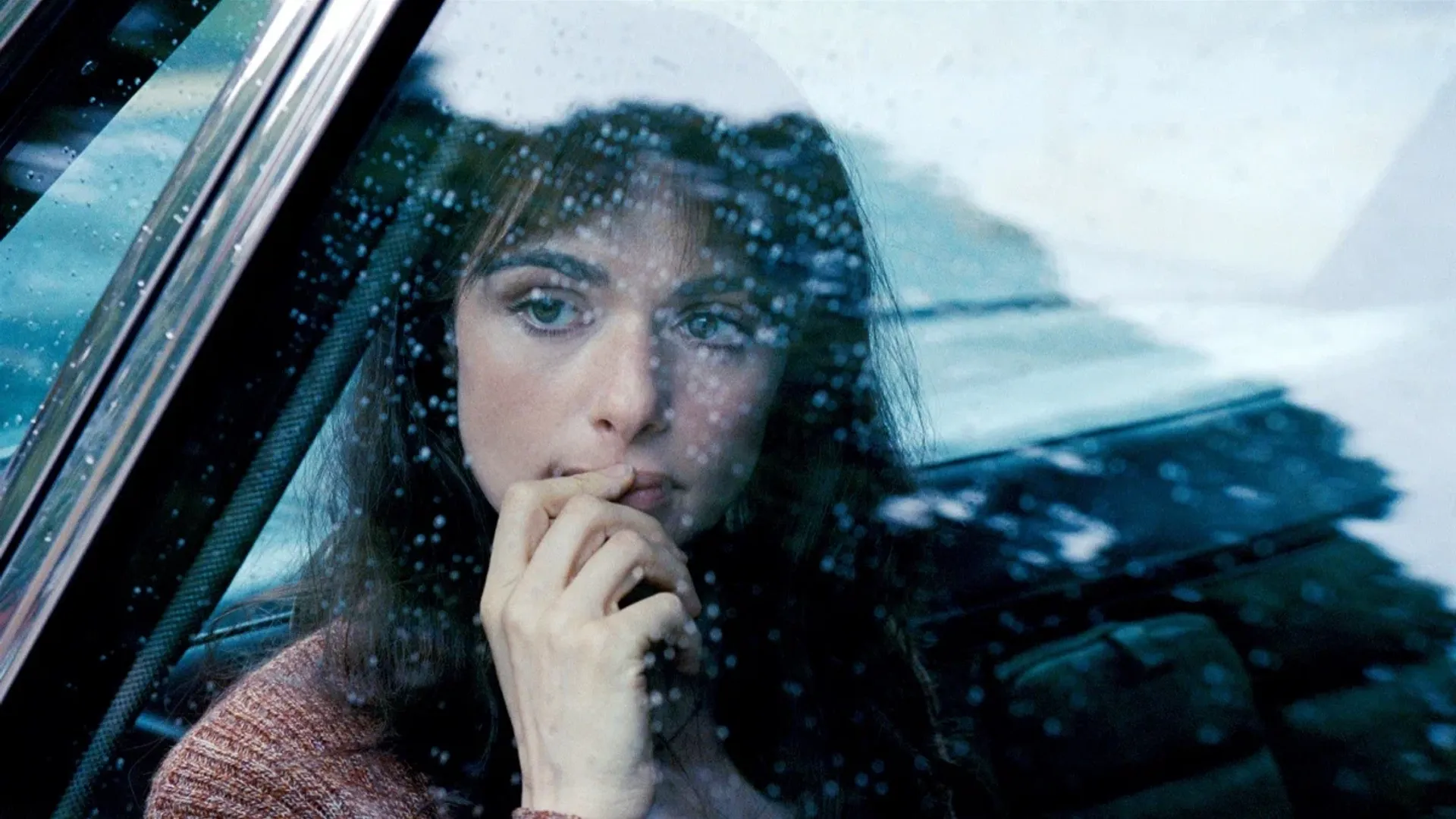 Close-up do rosto da atriz Rachel Weisz filmado através do vidro de um carro na chuva
