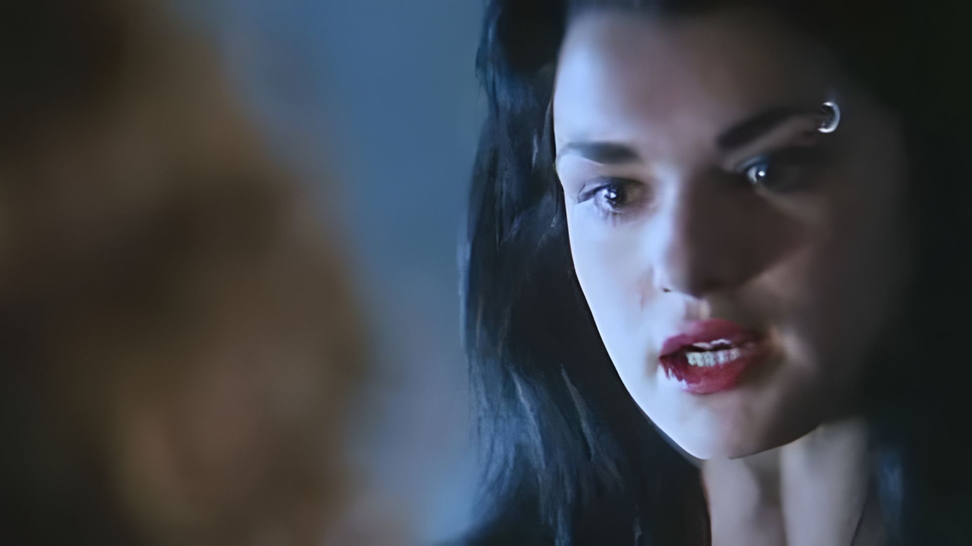 Um plano aproximado do rosto de Rachel Weisz, tem batom vermelho vivo e um piercing na sobrancelha