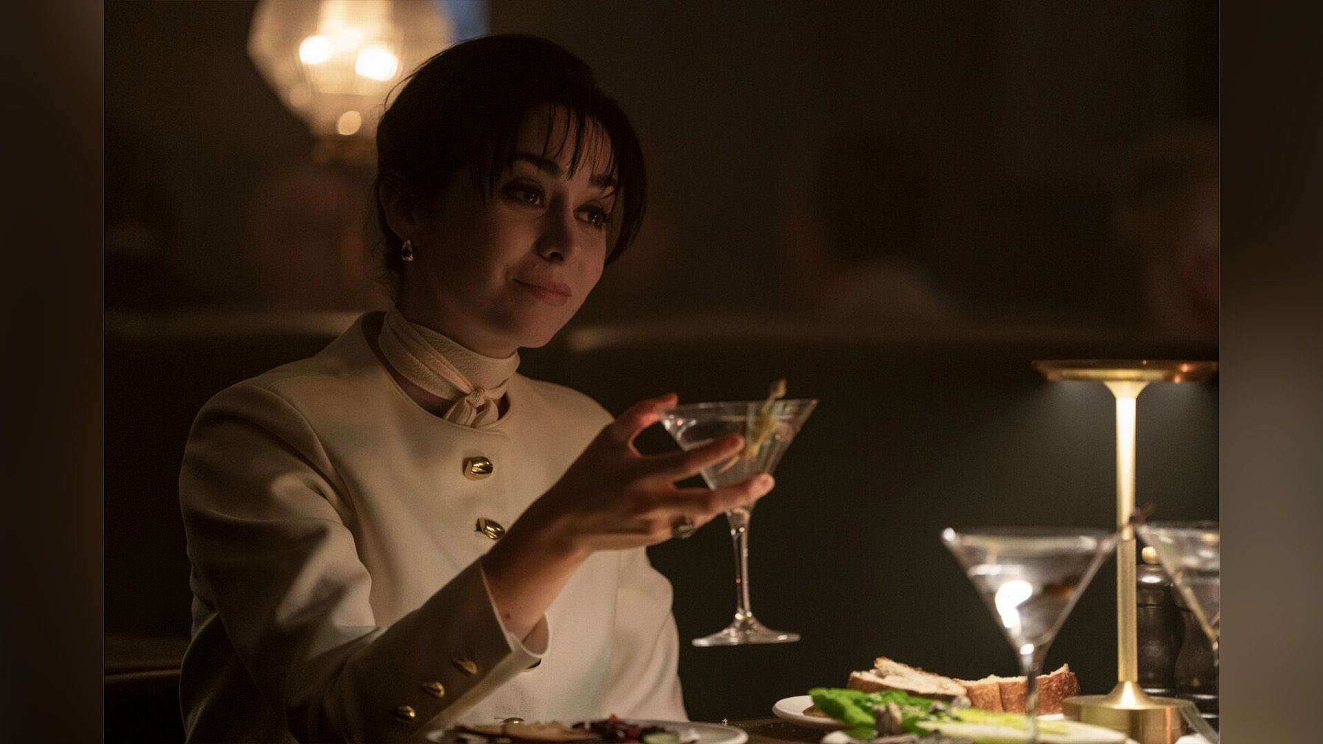 Cristin Milioti de fato branco segurando um copo numa cena da série "The Penguin"