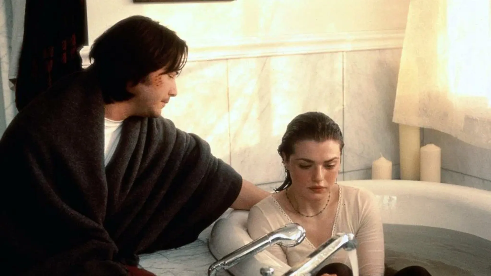 A jovem Rachel Weisz está sentada na banheira vestida, ao lado dela está sentado o jovem Keanu Reeves