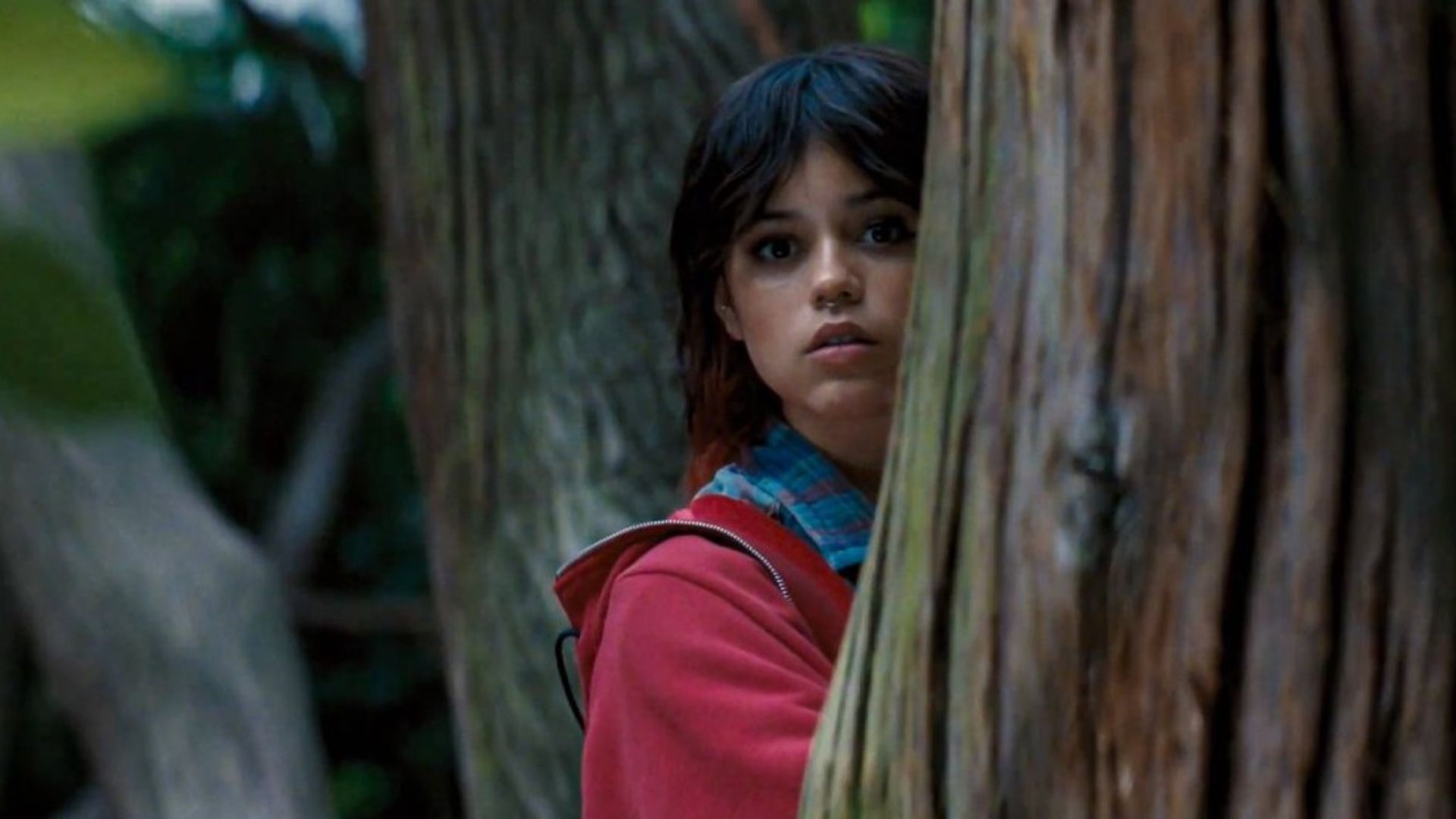A atriz Jenna Ortega espreita por detrás de uma árvore, vestindo uma camisola rosa