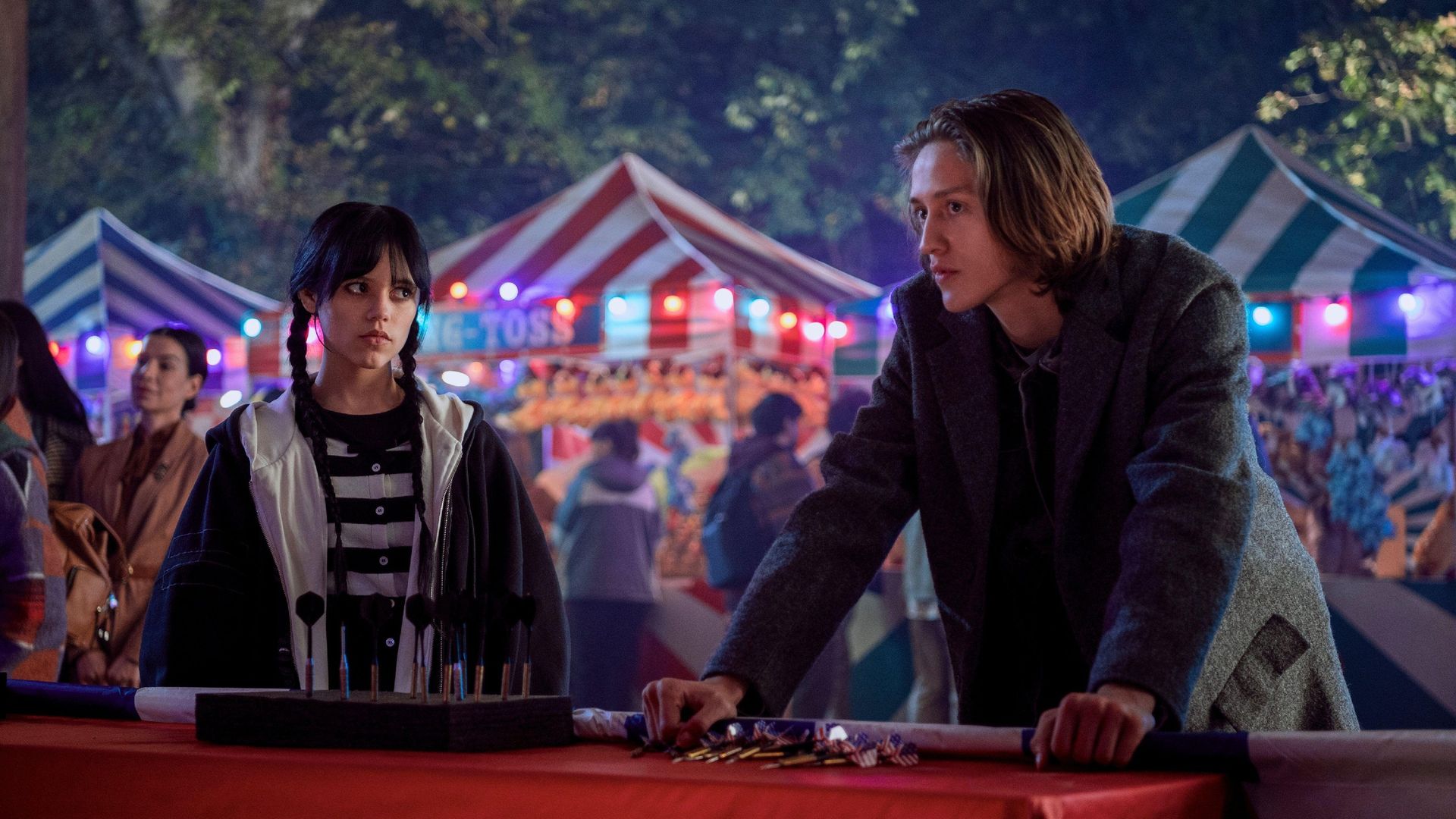 Jenna Ortega e Percy Hynes White em primeiro plano com uma feira ao fundo