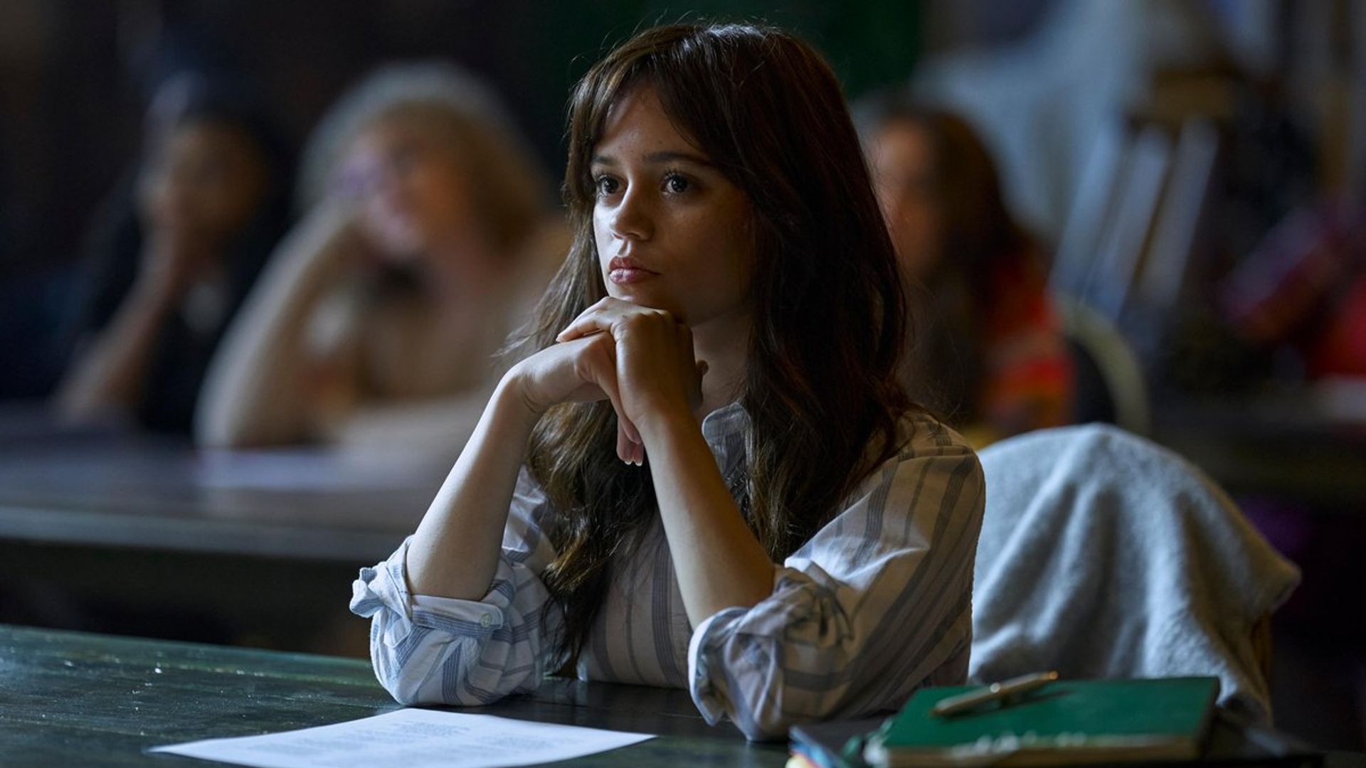 Jenna Ortega numa camisola branca sentada numa secretária durante a aula com uma expressão concentrada