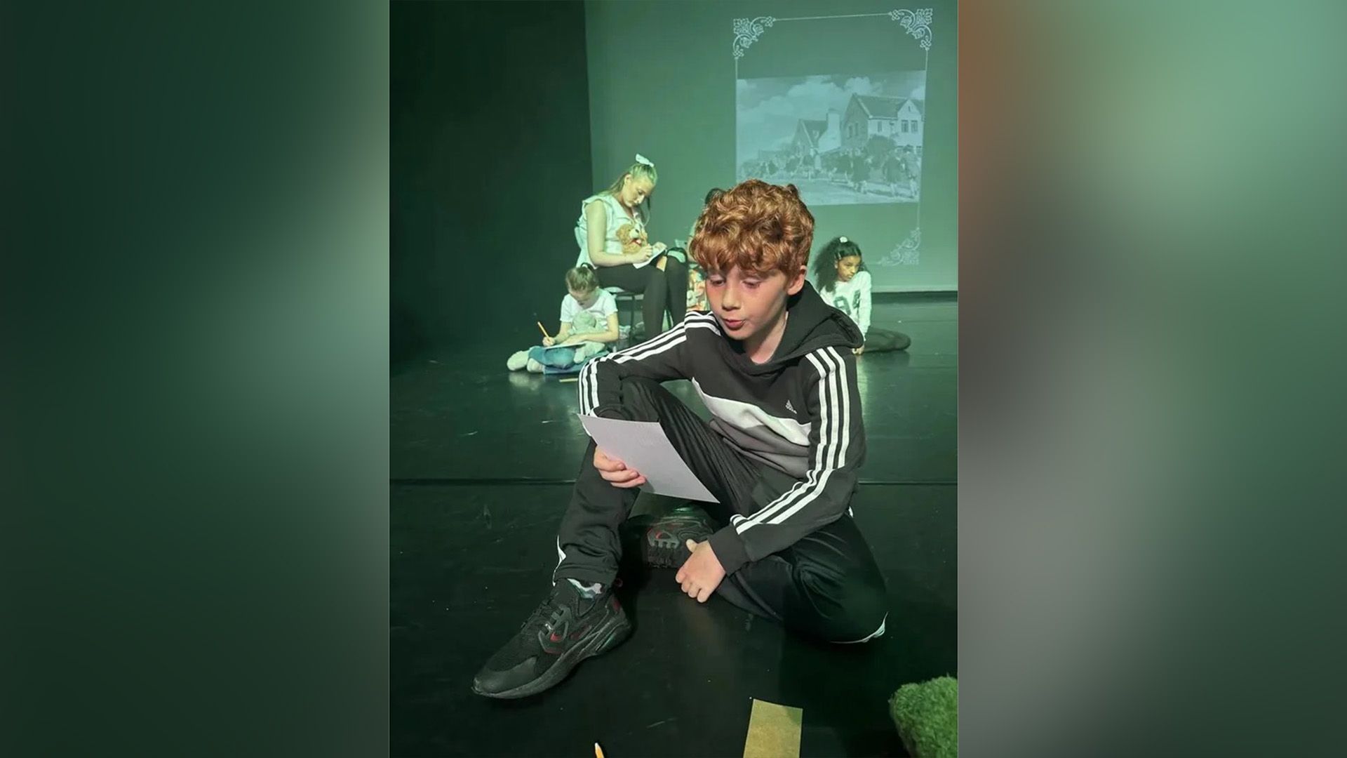 Alastair Stout com um fato de treino sentado num palco de teatro a ler de uma folha