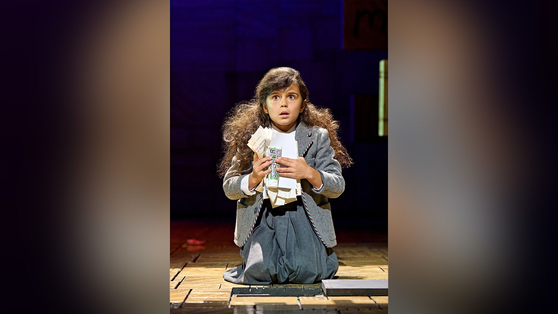 Arabella Stanton num vestido cinzento ajoelhada num palco de teatro, segurando folhas de papel amarrotadas nas mãos