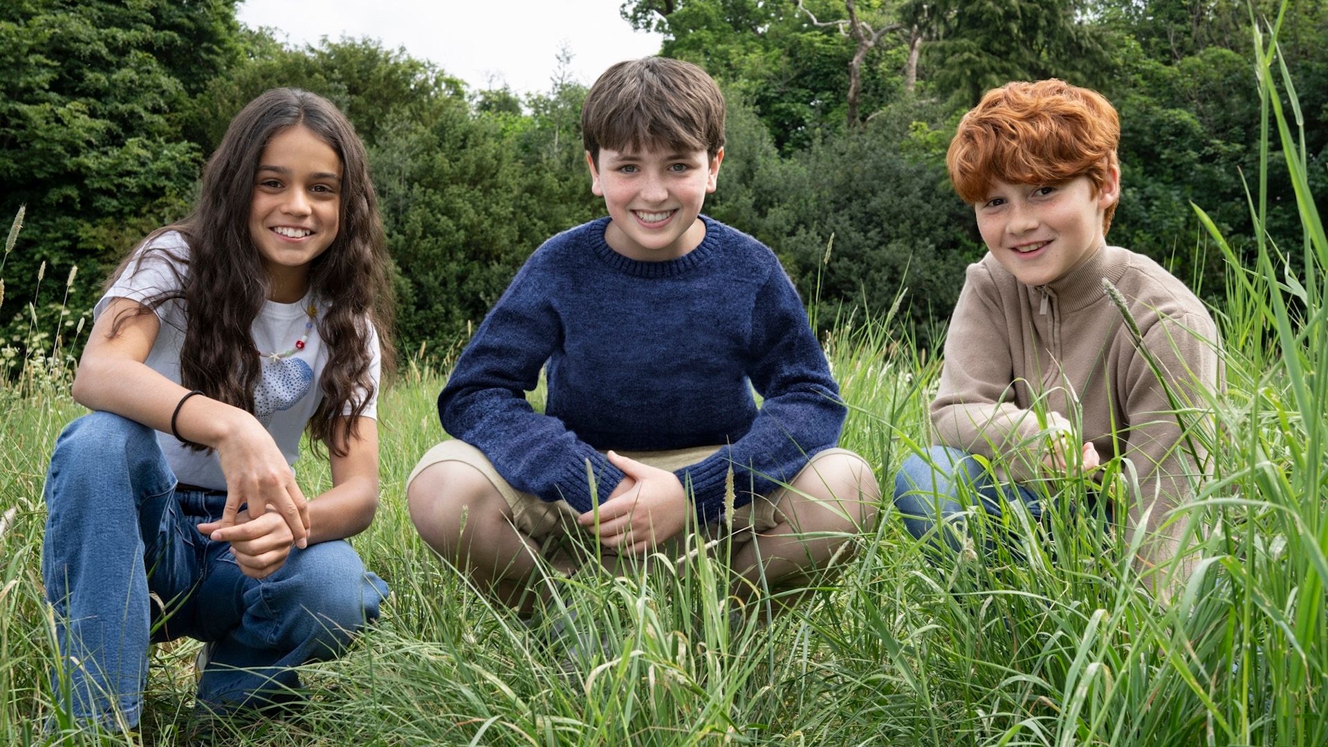 Os jovens atores Arabella Stanton, Dominic McLaughlin e Alastair Stout agachados na relva e sorrindo
