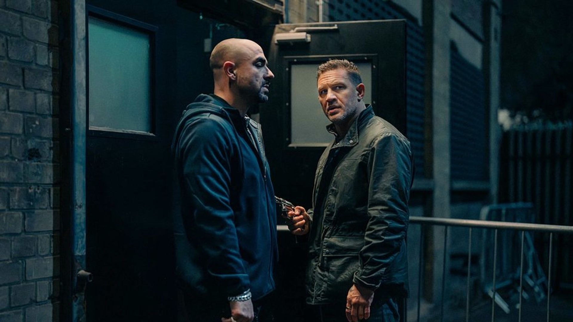 Tom Hardy e outro homem vestidos de escuro estão a conversar junto a uma saída preta durante a noite, um deles segura uma arma