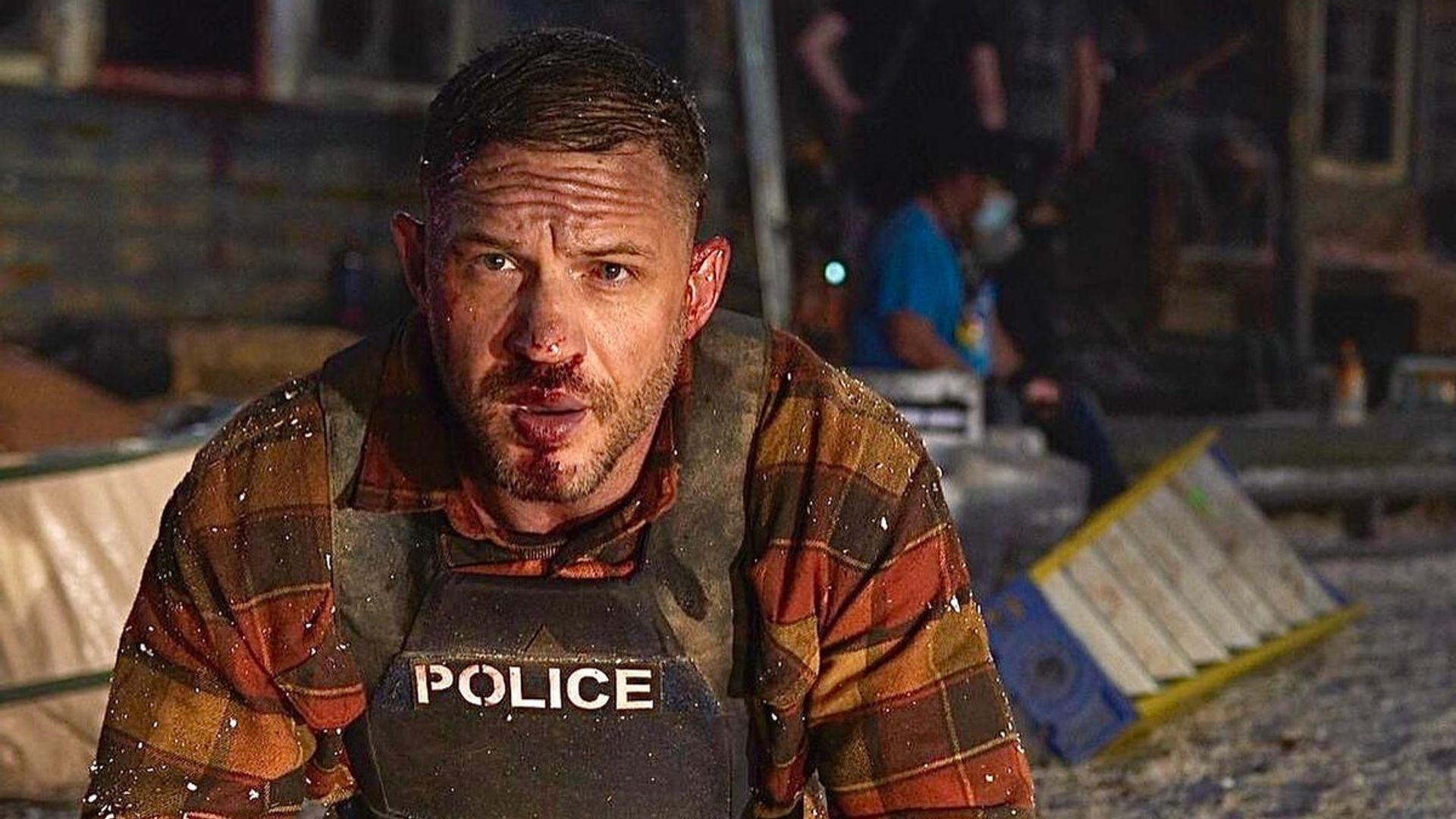 Tom Hardy num colete à prova de bala com o rosto machucado está ajoelhado no local do incidente