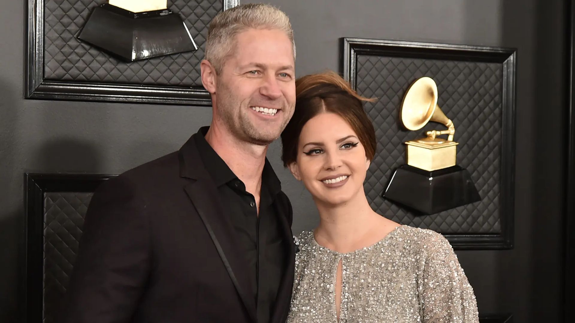 Lana Del Rey e Sean Larkin posam na passadeira vermelha dos Grammy Awards contra uma parede preta com imagens de gramofones dourados