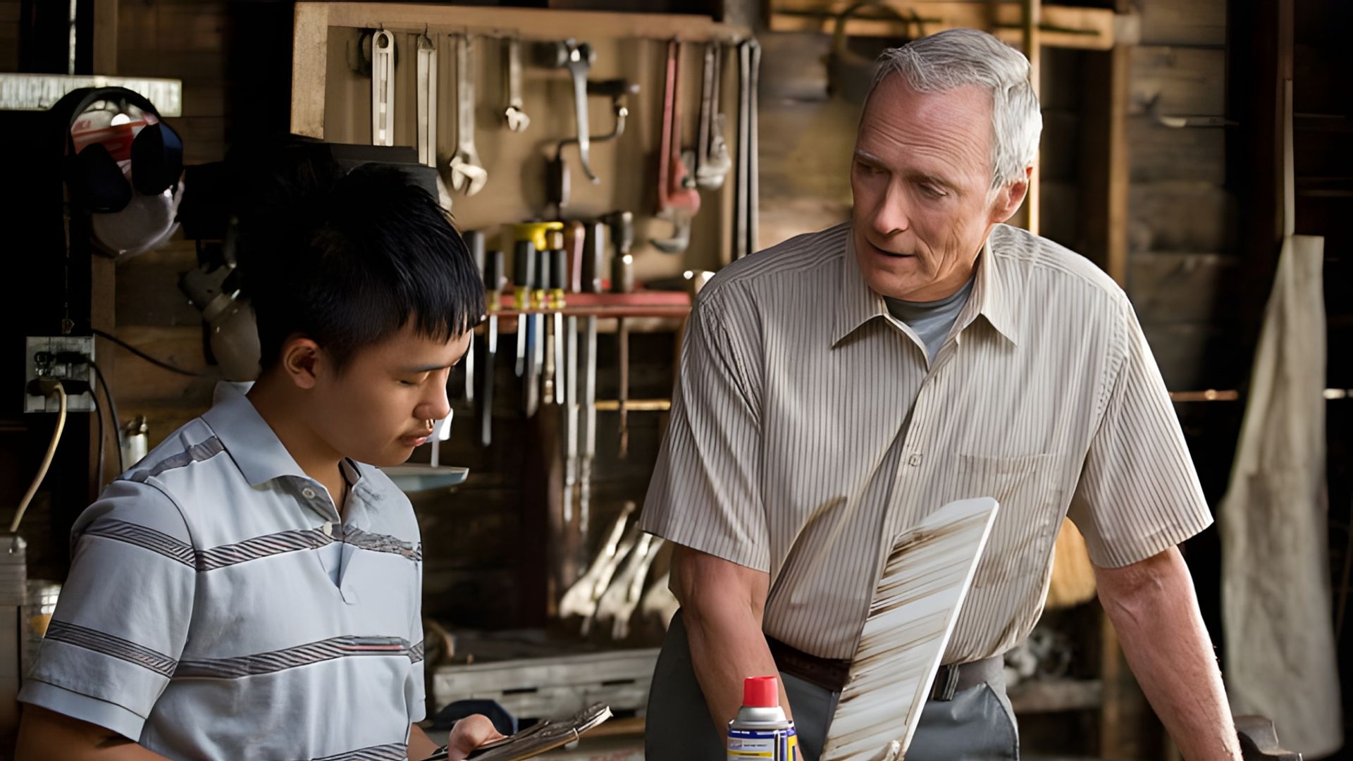 Clint Eastwood e um adolescente trabalham numa oficina, discutindo uma ferramenta contra o pano de fundo de uma bancada com ferramentas penduradas