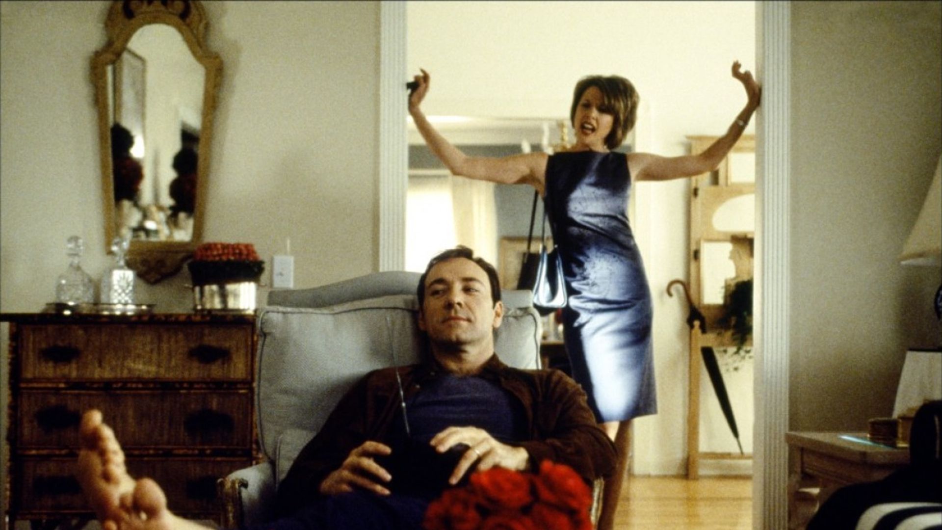 Kevin Spacey senta-se relaxado numa cadeira com um comando enquanto uma mulher em vestido de noite atrás dele gesticula emocionalmente na porta