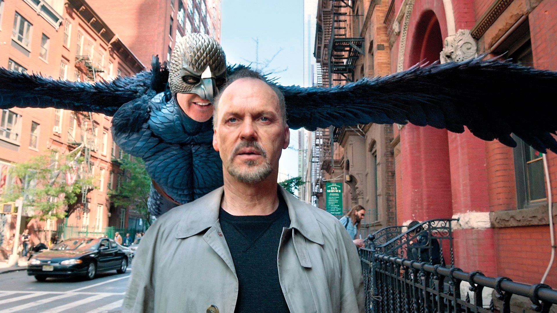 Michael Keaton caminha pela rua enquanto atrás dele paira uma personagem fantástica com asas e uma máscara de pássaro