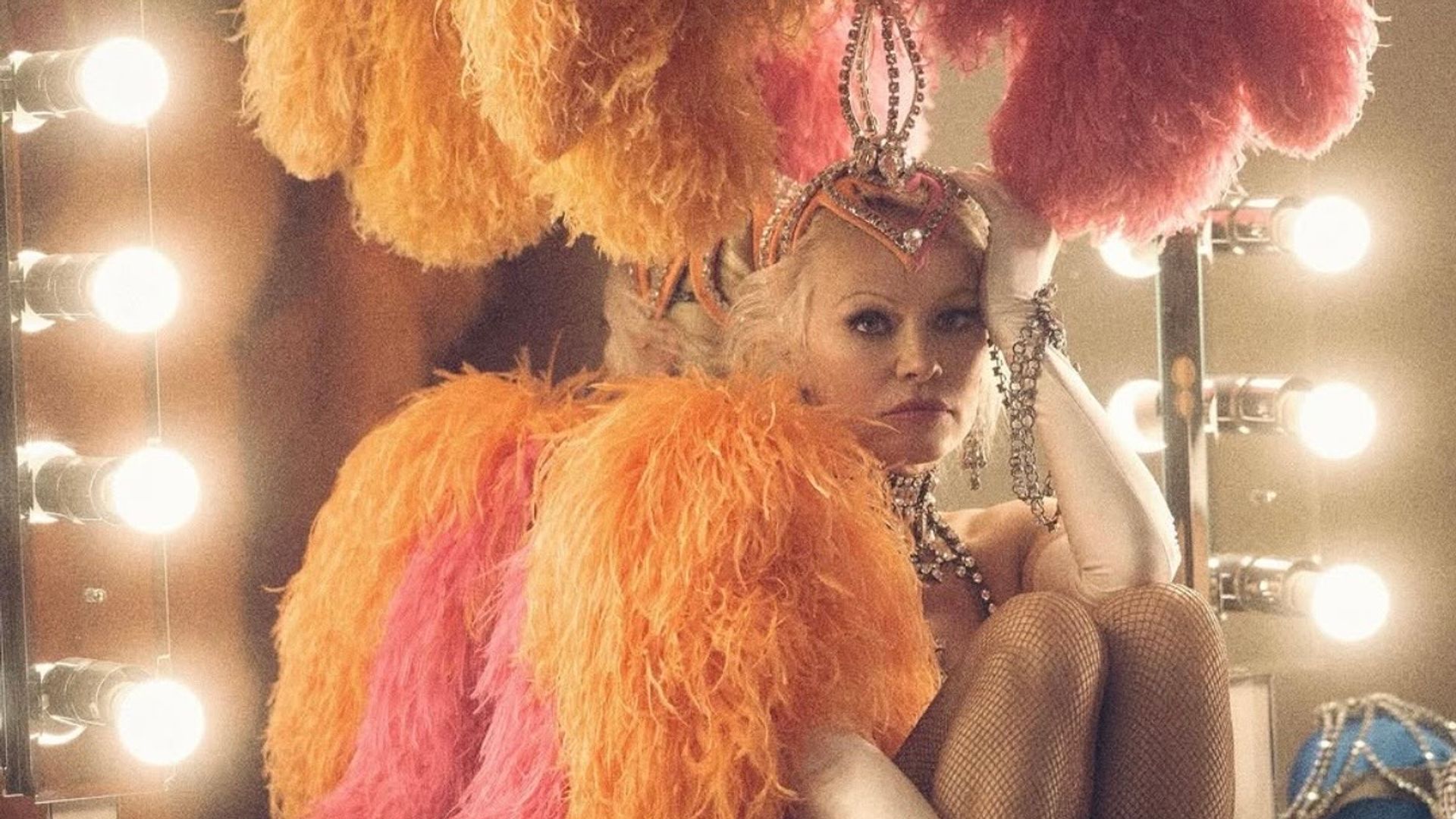 Pamela Anderson num traje de espetáculo cintilante com penas e adornos está sentada ao espelho do camarim, apoiando pensativamente a cabeça na mão