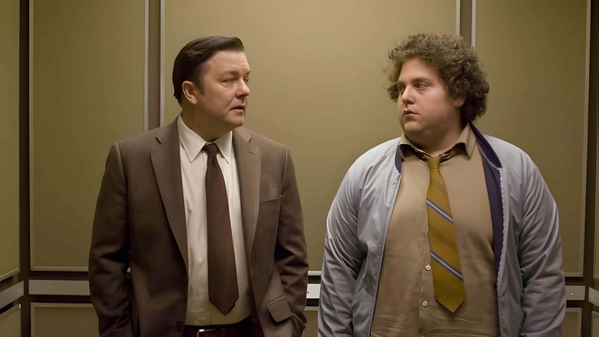 Ricky Gervais e Jonah Hill em traje de negócios de pé num elevador, olhando um para o outro com expressões sérias nos rostos