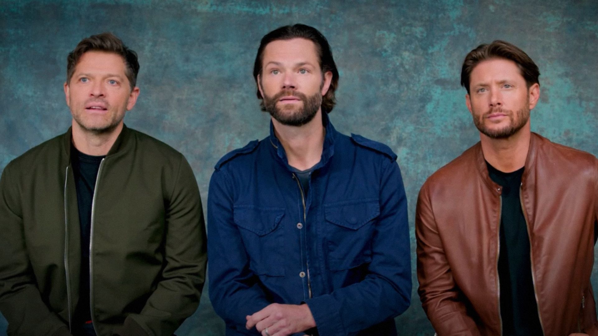 Os atores Misha Collins, Jared Padalecki e Jensen Ackles sentam-se contra um fundo azul usando casacos coloridos