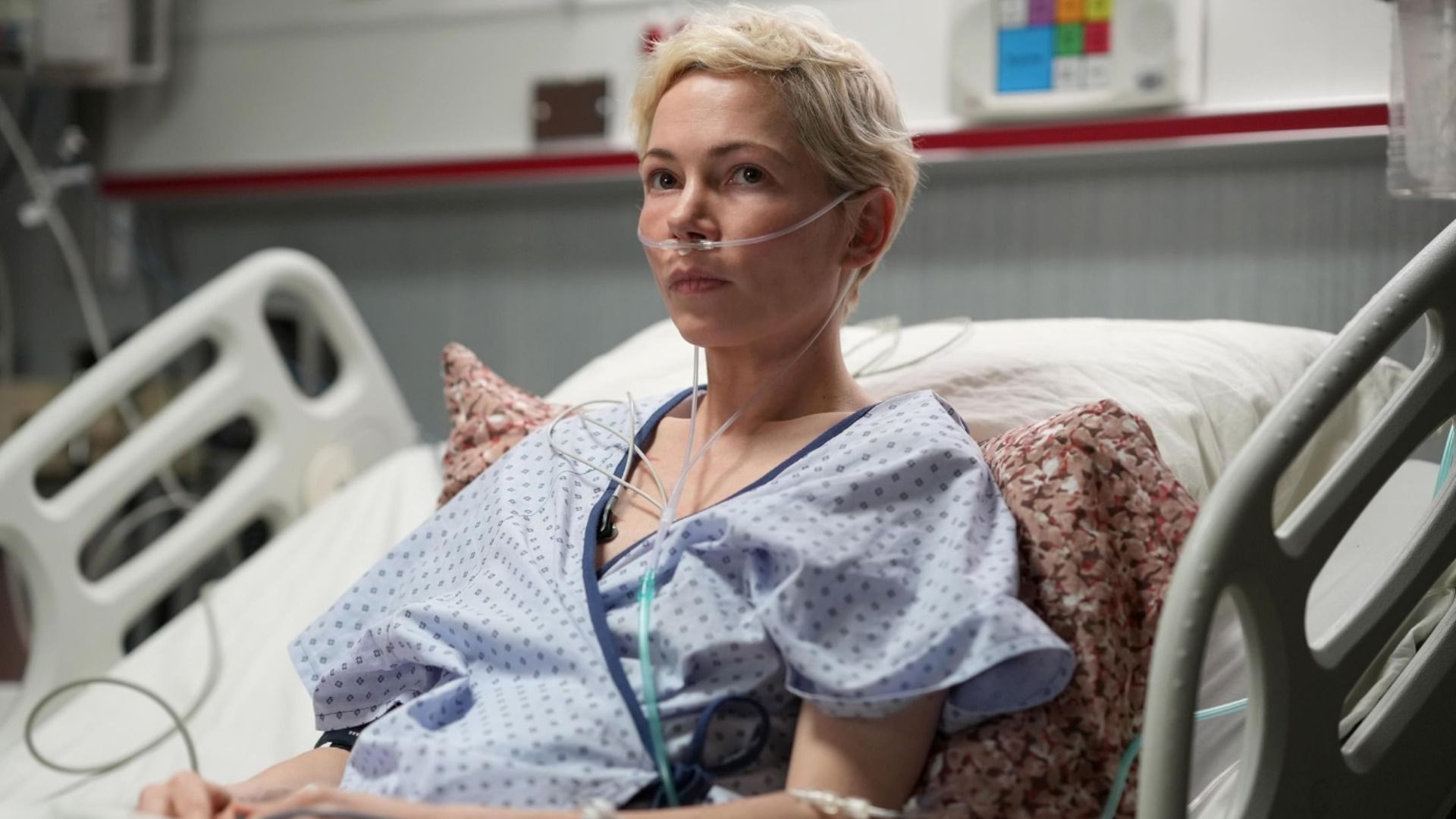 A atriz Michelle Williams com roupa de paciente hospitalar deitada numa cama com um tubo no nariz