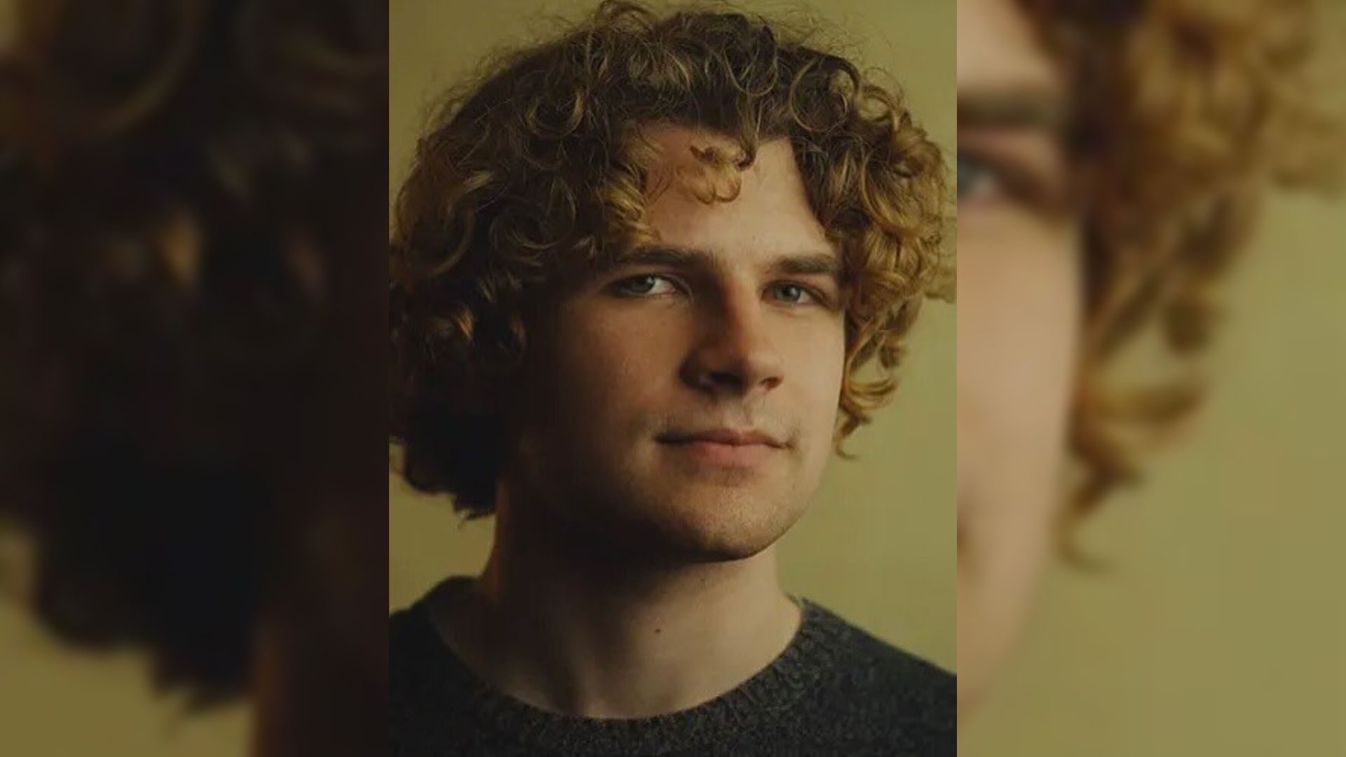 Um jovem de cabelo encaracolado com uma camisola preta está contra um fundo amarelo