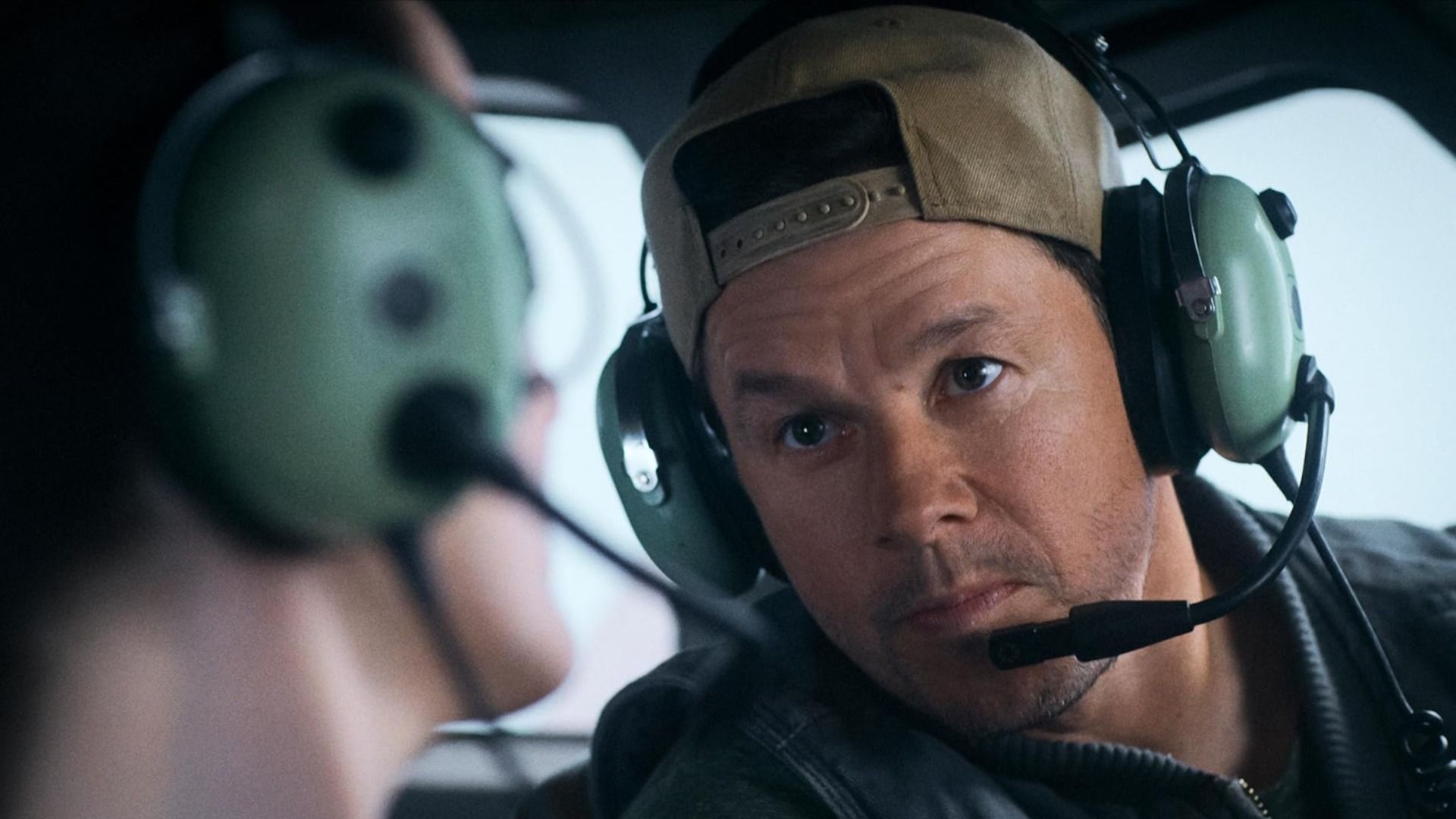 O ator Mark Wahlberg usando um boné e auscultadores com microfone sentado num avião